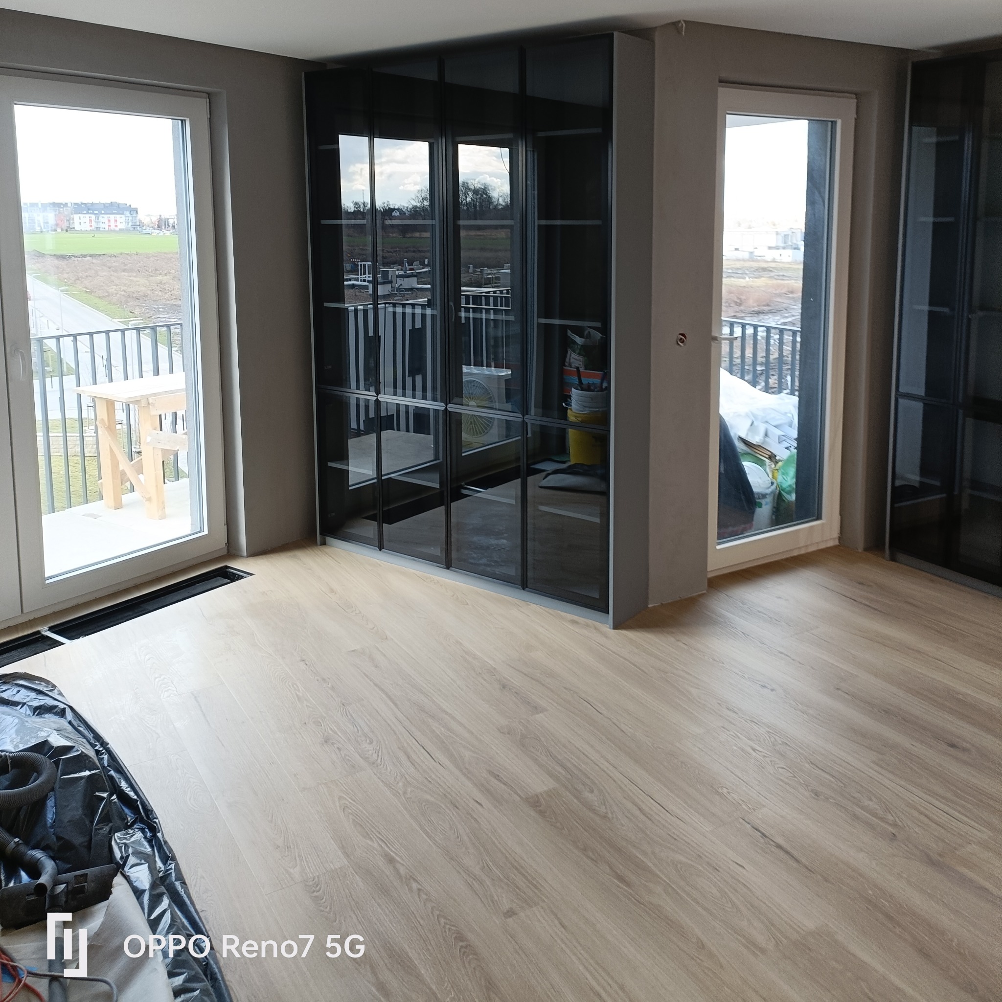 Montaż podłogi laminowanej w jasnym odcieniu drewna, widoczne okna balkonowe z widokiem na zewnątrz i zabudowa meblowa z czarnymi, przeszklonymi frontami.