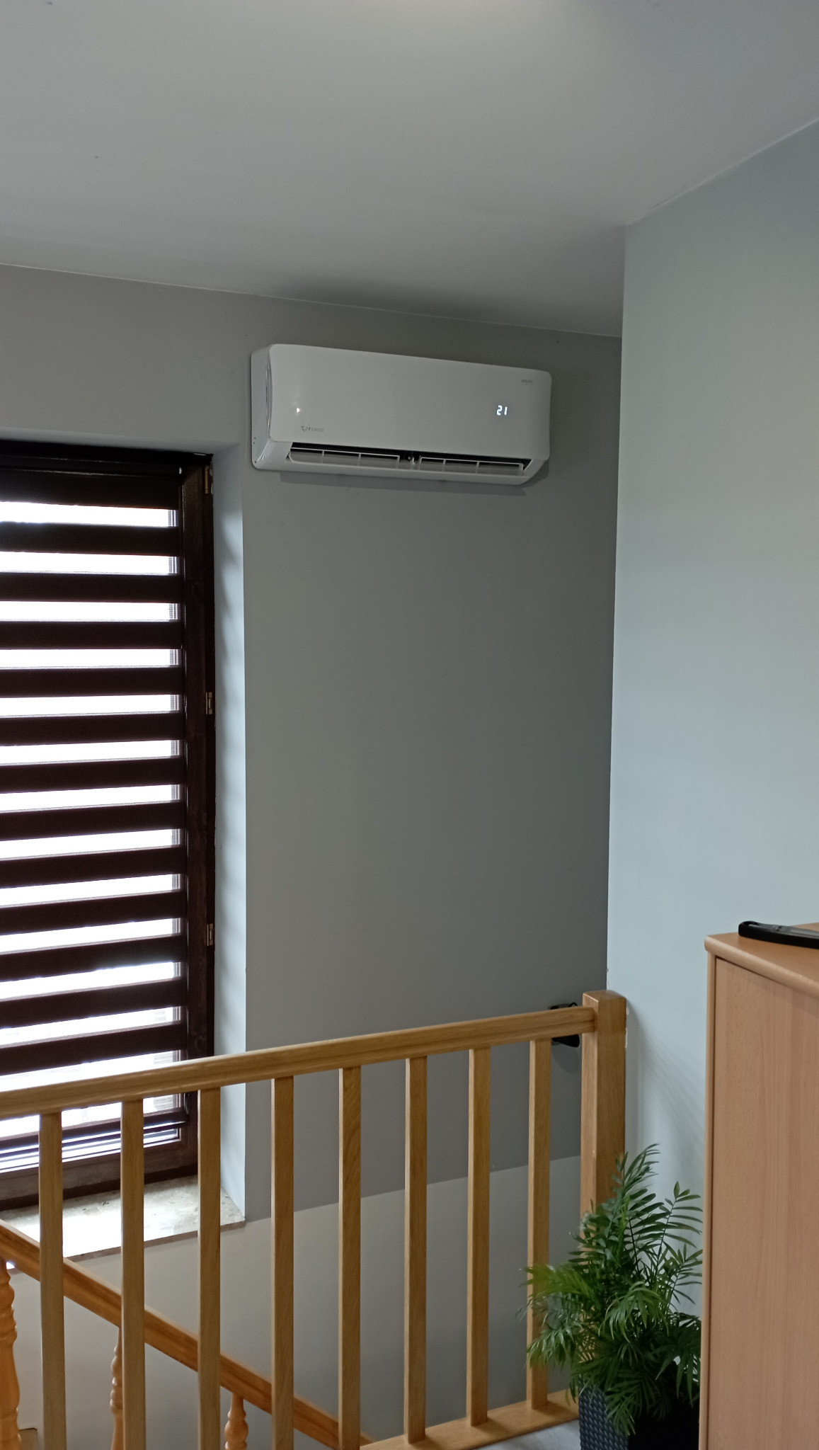 Biały klimatyzator ścienny zamontowany w szarym pokoju, widoczny zza drewnianej balustrady schodów i rośliny doniczkowej.