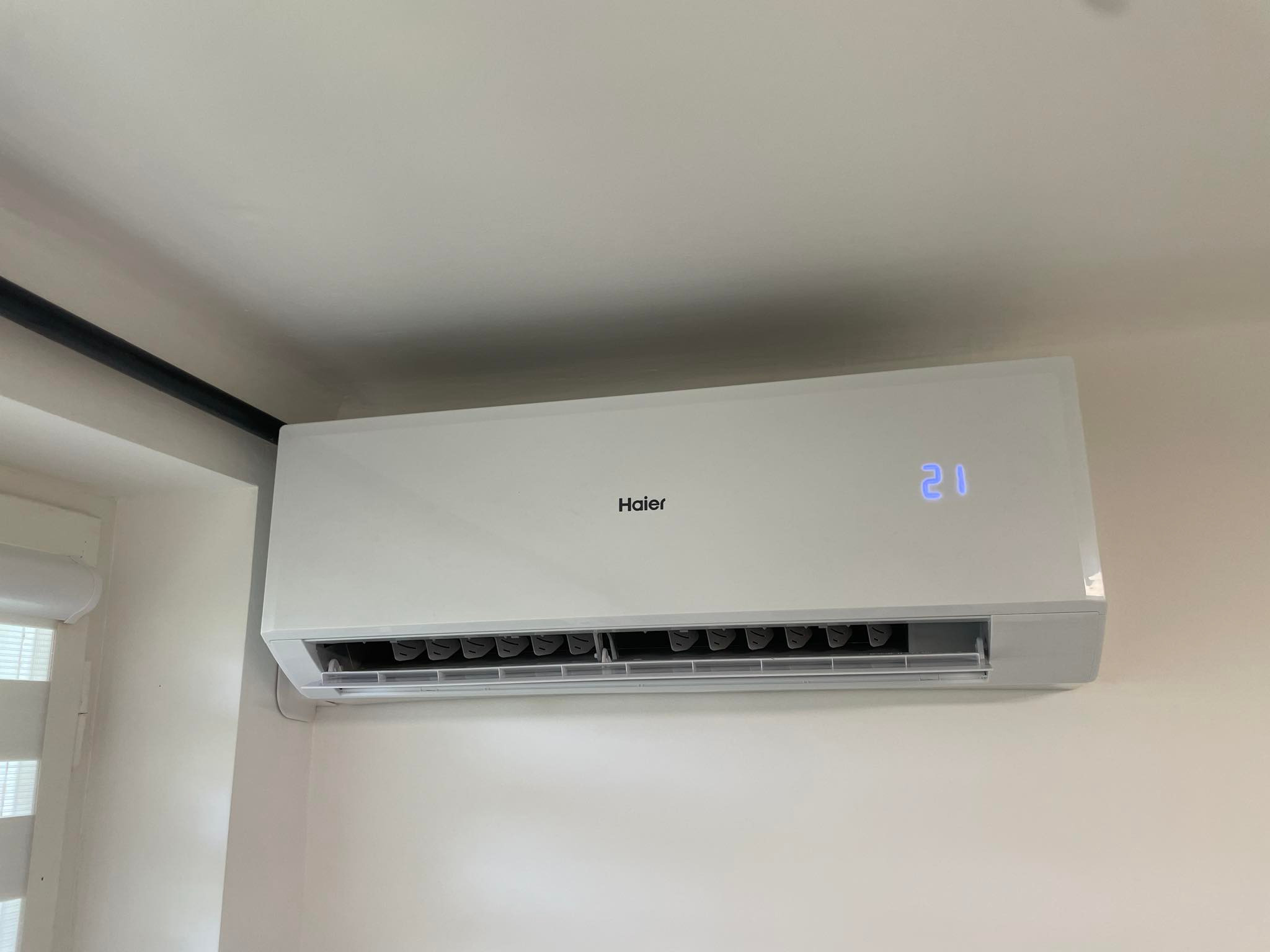 Biała jednostka klimatyzacji Haier zamontowana na ścianie, wyświetlająca temperaturę 21 stopni Celsjusza, widoczna część sufitu i rolety okienne w tle.