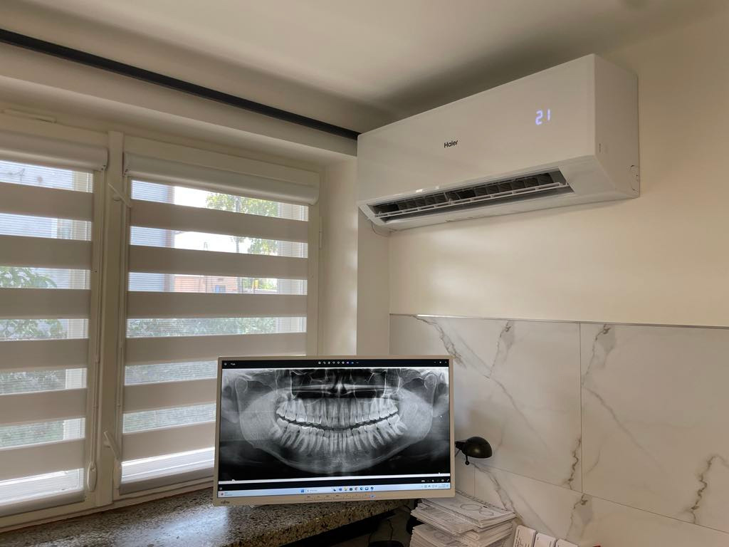 Biały klimatyzator Haier zamontowany na ścianie w pomieszczeniu, z widocznym wyświetlaczem temperatury. Na pierwszym planie monitor wyświetlający zdjęcie rentgenowskie szczęki, obok okno z żaluzjami.