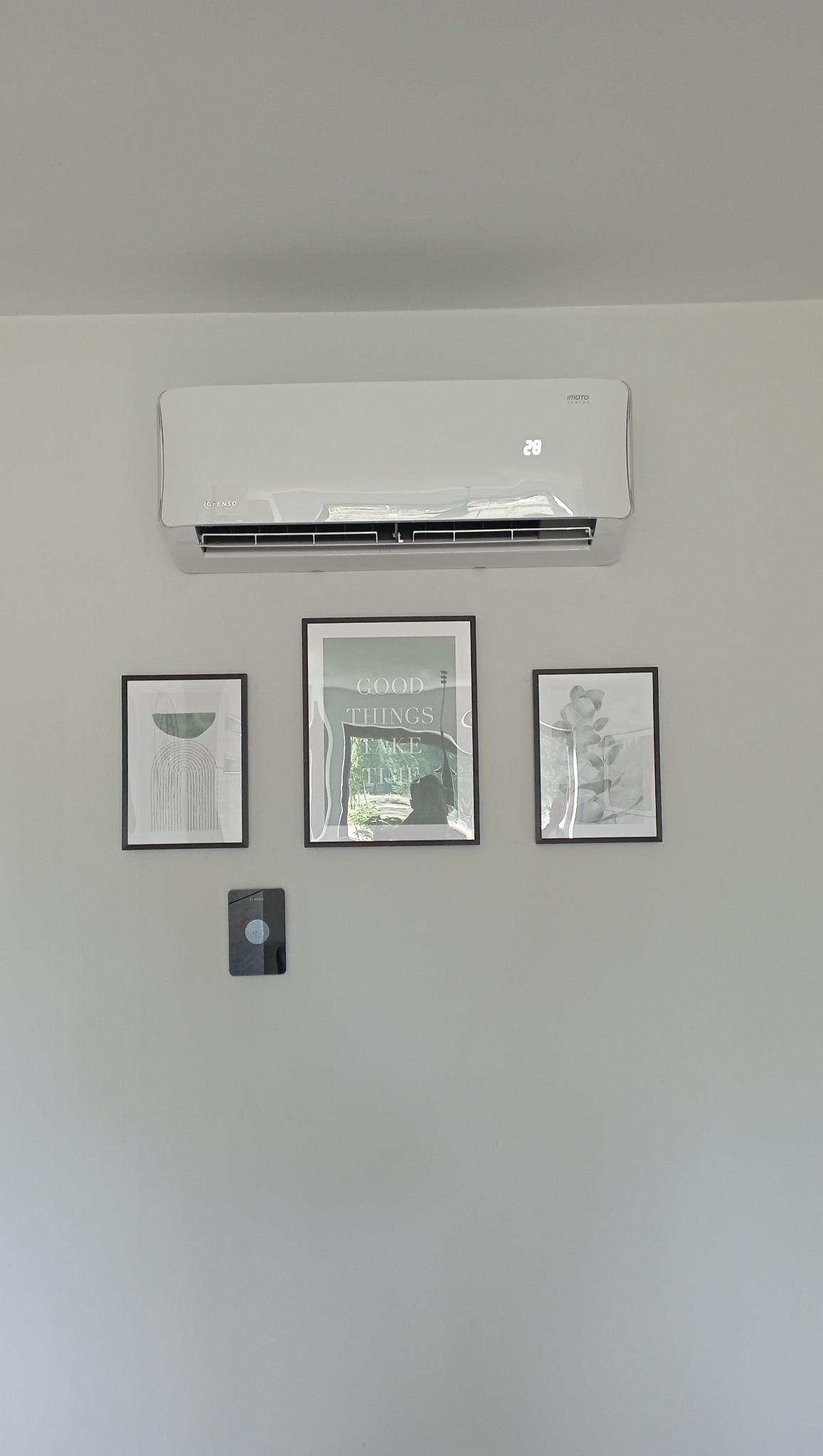 Biały klimatyzator ścienny marki Rotenso zamontowany w pomieszczeniu, z wyświetlaczem wskazującym temperaturę 28 stopni Celsjusza, poniżej trzy grafiki w czarnych ramkach i czujnik.
