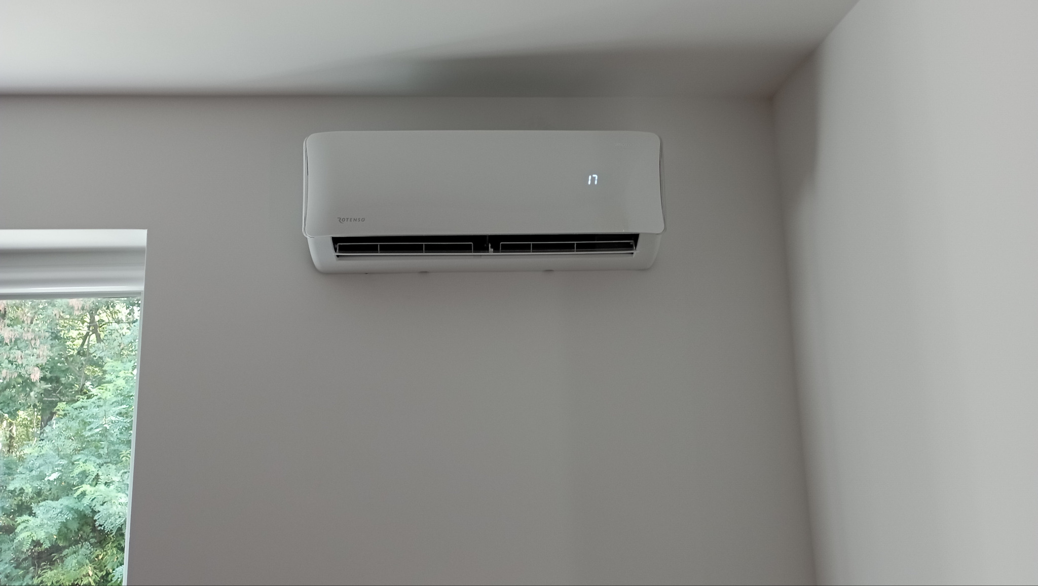Biała klimatyzacja Rotenso zamontowana na szarej ścianie w pokoju, z widocznym oknem i zielenią na zewnątrz.