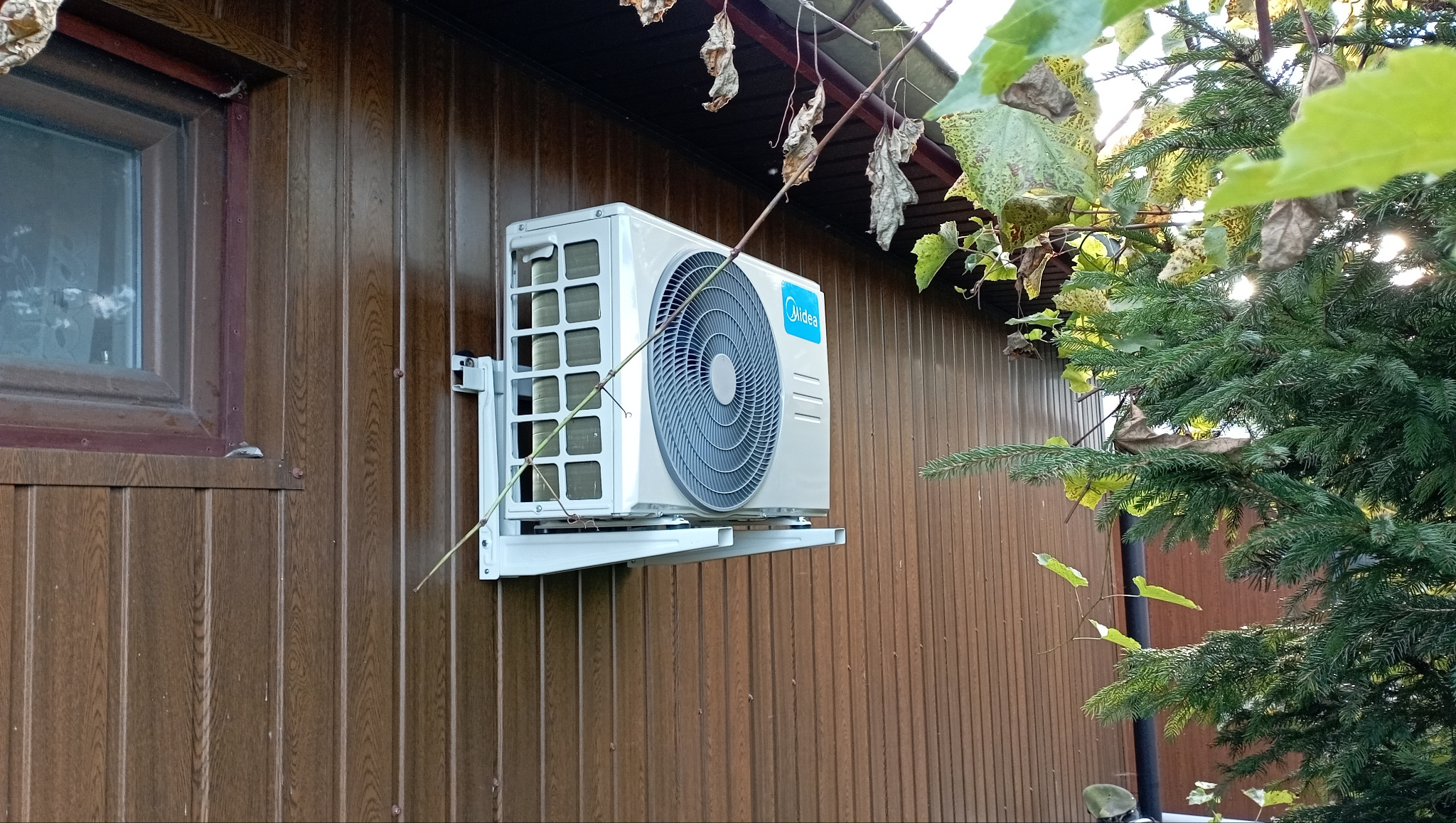 Zewnętrzna jednostka klimatyzatora Midea zamontowana na brązowej, drewnianej ścianie budynku, widoczna z boku, z zielenią w tle.
