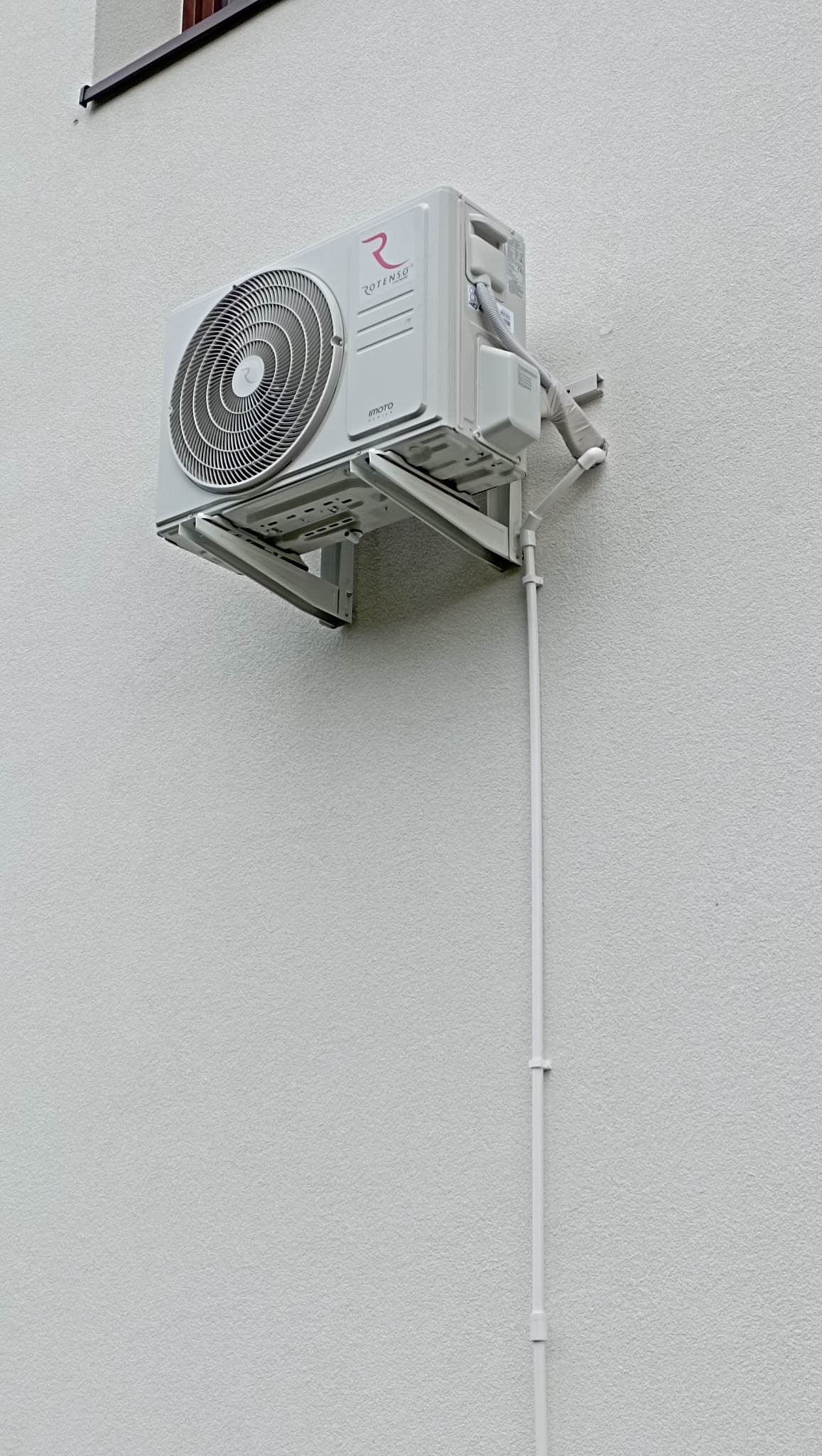 Zewnętrzna jednostka klimatyzacji Rotenso zamontowana na białej, teksturowanej ścianie budynku, z widocznymi rurami odprowadzającymi skropliny i elementami mocującymi.