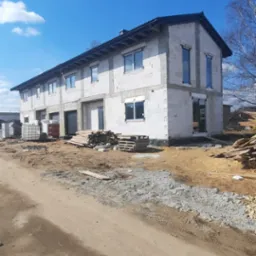 Budynek w stanie surowym otwartym z zamontowanymi oknami i drzwiami, widoczne materiały budowlane i plac budowy.