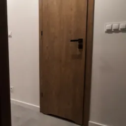 Zamknięte drzwi wewnętrzne z drewnopodobnym wykończeniem i czarną klamką, widoczne na tle białej ściany i szarej podłogi.