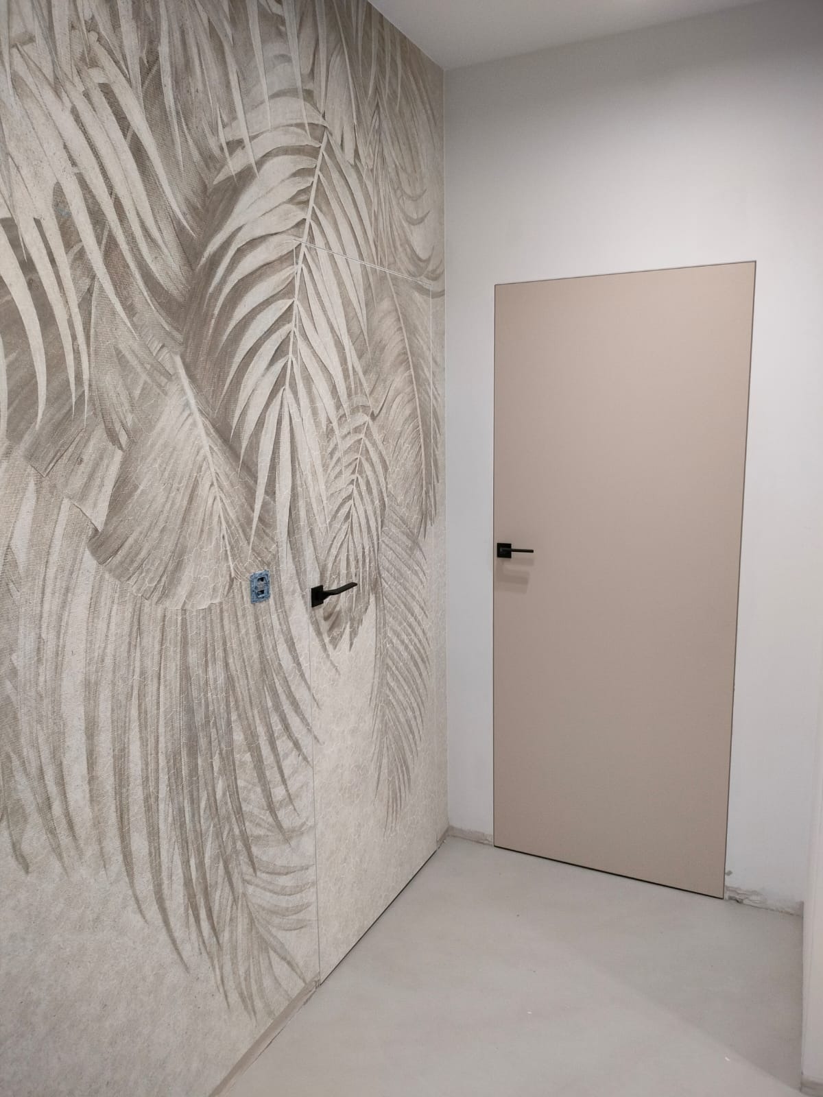 Beżowe drzwi wewnętrzne z czarną klamką, zamontowane w pomieszczeniu z tapetą w motywie liści palmowych, widoczne gniazdko elektryczne.