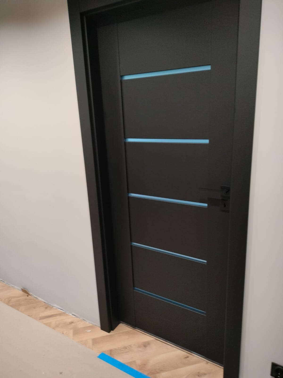 Ciemne, nowoczesne drzwi wewnętrzne z poziomymi, błękitnymi wstawkami, zamontowane w jasnym pomieszczeniu z parkietem w jodełkę, zabezpieczone niebieską taśmą malarską u dołu.
