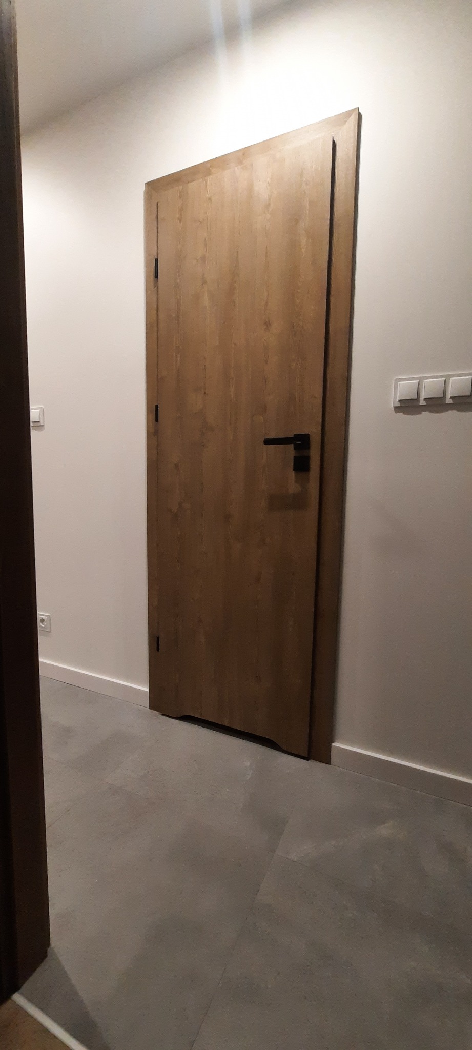 Zamknięte drzwi wewnętrzne z drewnopodobnym wykończeniem i czarną klamką, widoczne na tle białej ściany i szarej podłogi.