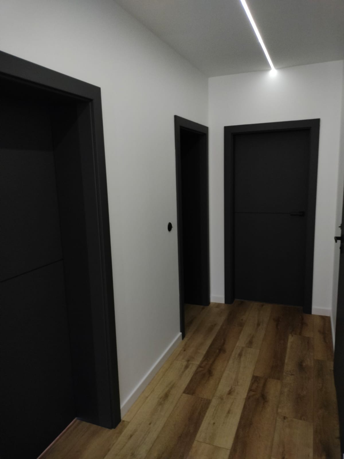 Wnętrze korytarza z trzema czarnymi drzwiami wewnętrznymi, podłoga z paneli drewnopodobnych, oświetlenie LED w suficie.