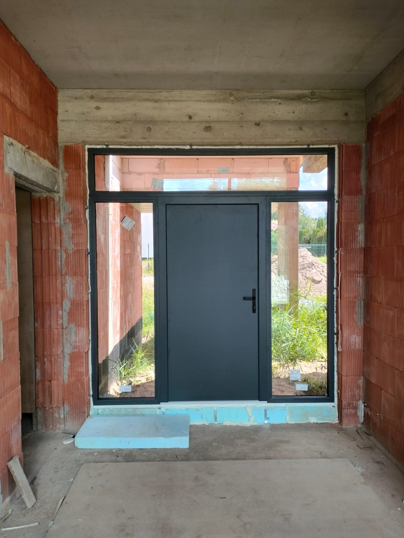 Ciemnoszare drzwi z przeszkleniami bocznymi i górnym, zamontowane w surowym stanie budynku z czerwonej cegły. Widoczne elementy montażowe i tymczasowe podesty z niebieskiego styropianu.