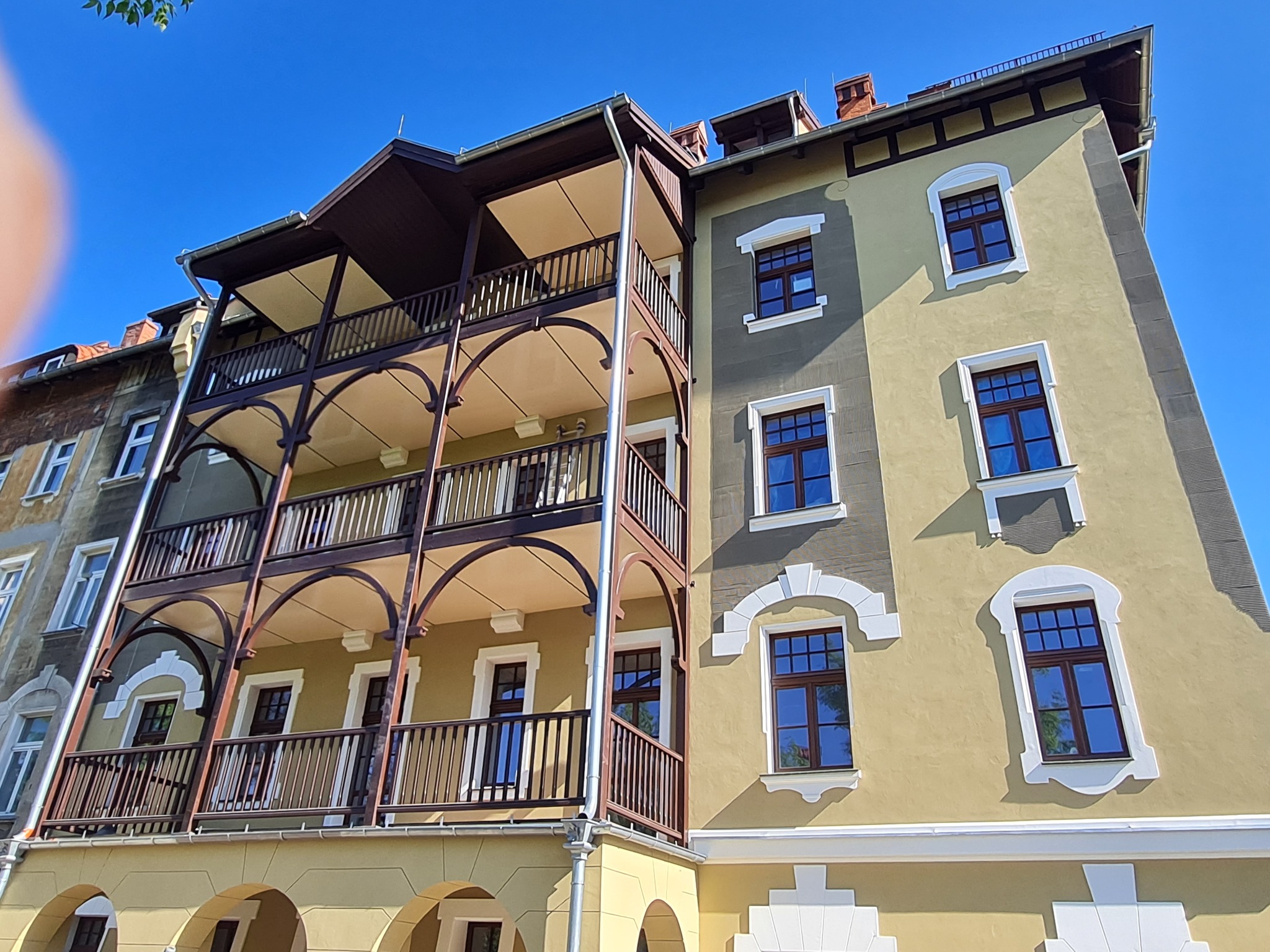 Odnowiona fasada kamienicy z balkonami, widok z dołu na żółto-szarą elewację z drewnianymi oknami i balustradami balkonów, błękitne niebo w tle.