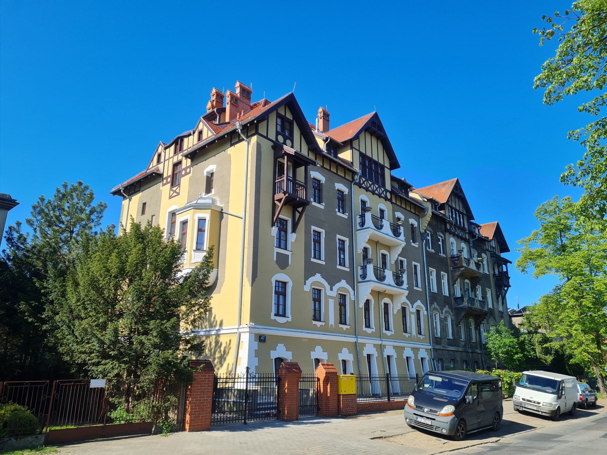 Odnowiona kamienica w stylu pruskim z balkonami, żółto-szara fasada, czerwona dachówka, błękitne niebo. Widok z dołu, auta zaparkowane na ulicy.