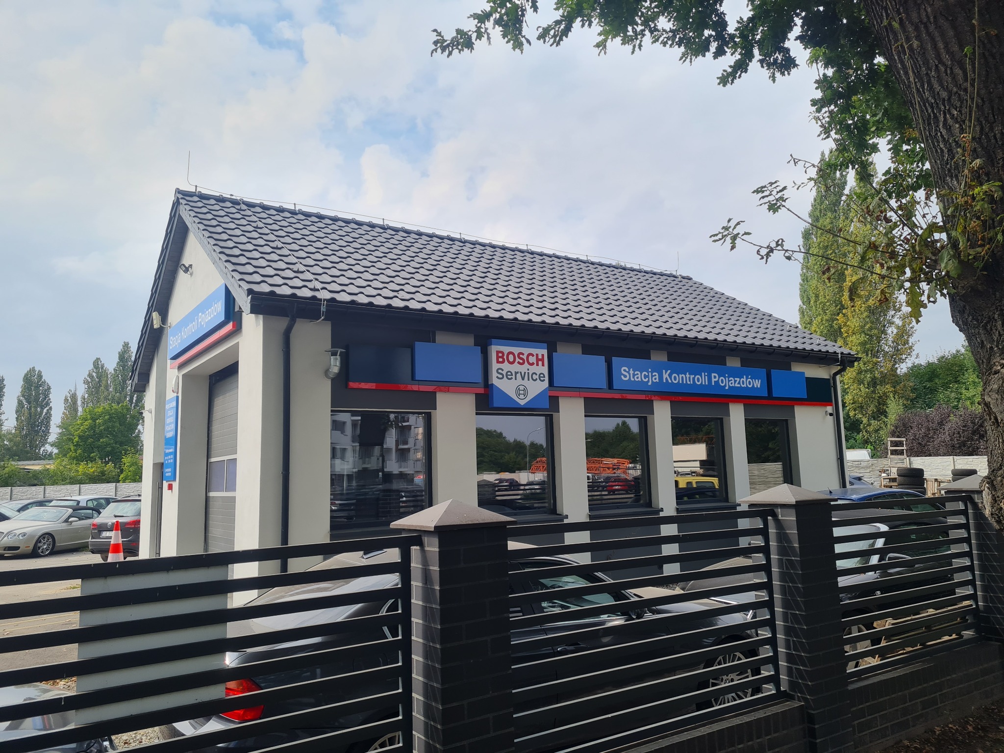 Budynek stacji kontroli pojazdów Bosch Service z szarą dachówką, widoczny zza czarnego ogrodzenia. Na parkingu zaparkowane samochody.