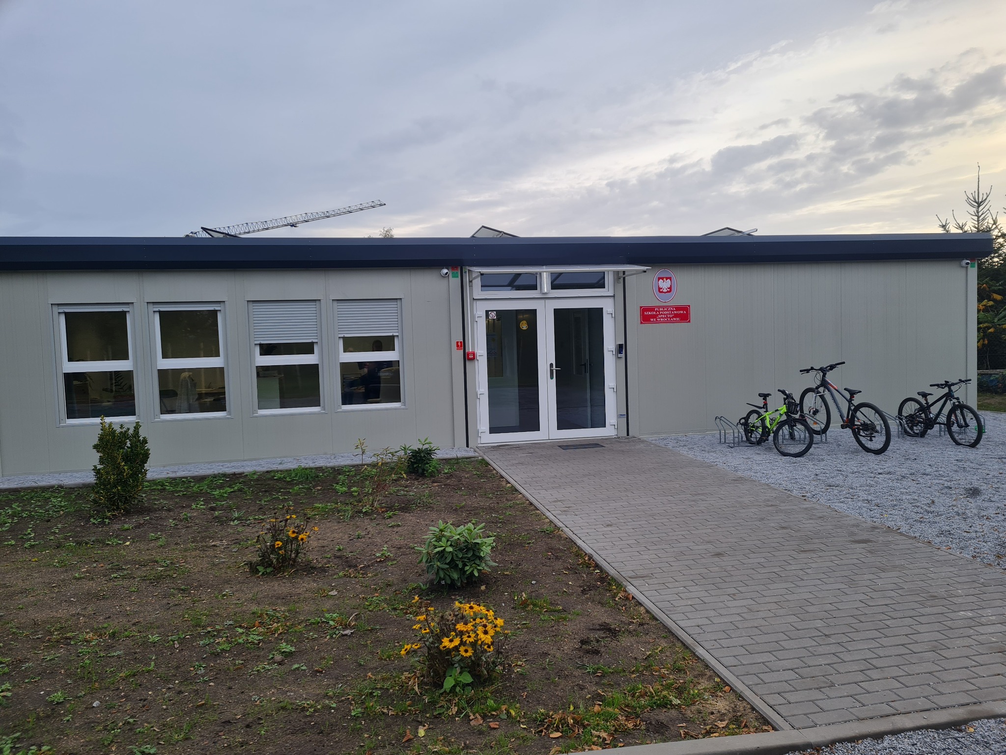 Modułowy budynek szkolny we Wrocławiu z rowerami przed wejściem. Widoczny fragment żurawia budowlanego w tle. Budynek parterowy z oknami i szarym wykończeniem.