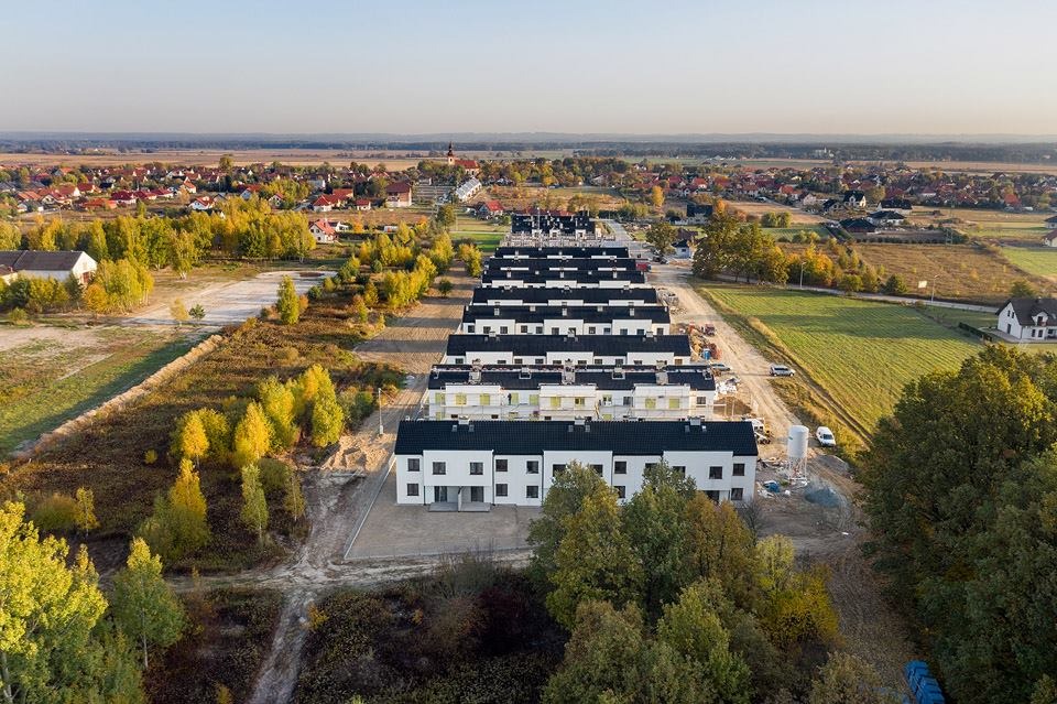 Widok z drona na szereg nowo wybudowanych, białych domów z czarnymi dachami, równo ustawionych na tle jesiennego krajobrazu. W tle widoczne zabudowania wsi.