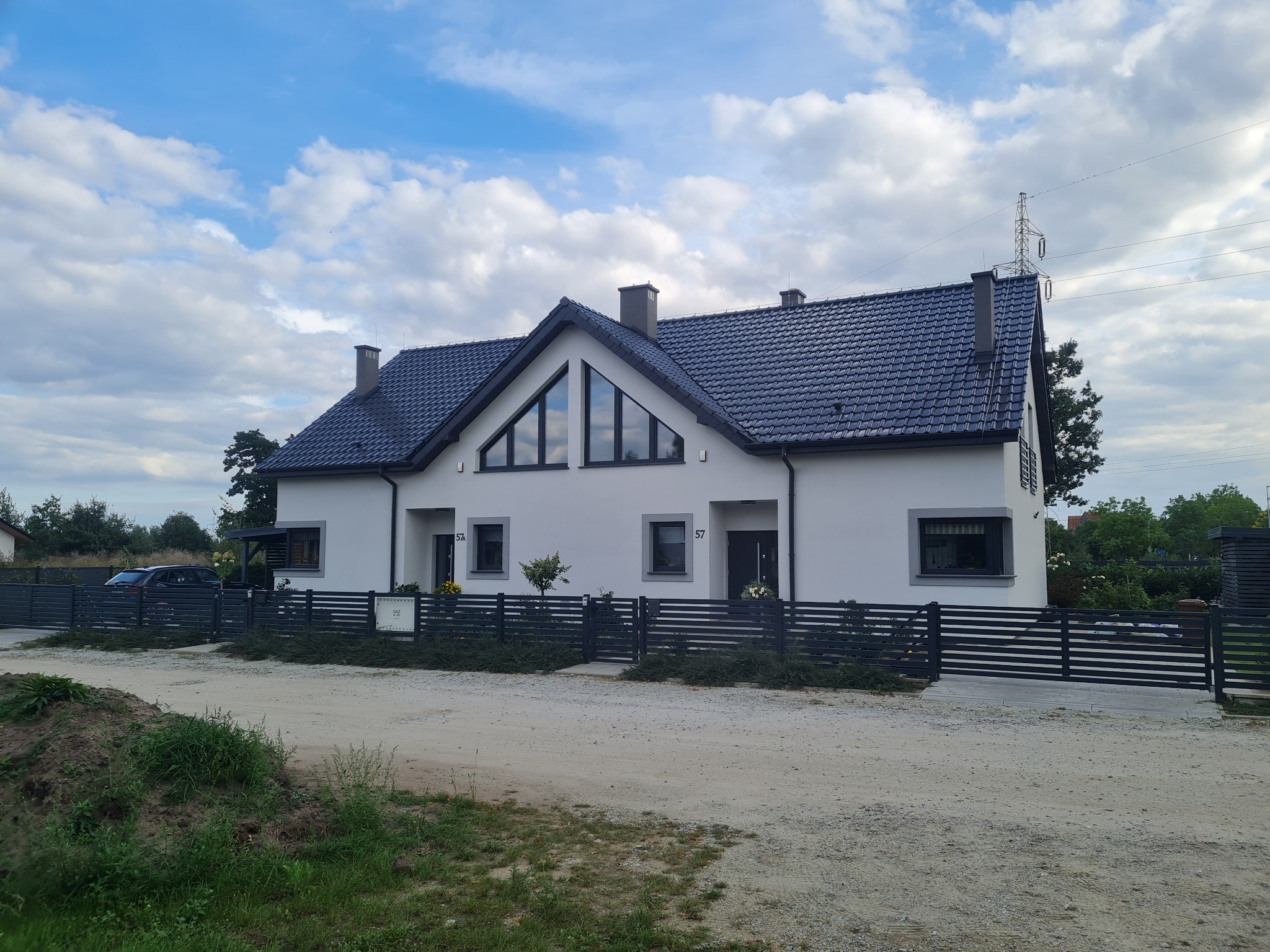 Nowoczesny, bliźniaczy dom z czarną dachówką i grafitowym ogrodzeniem, numer 57 na każdym segmencie, błękitne niebo z obłokami w tle.