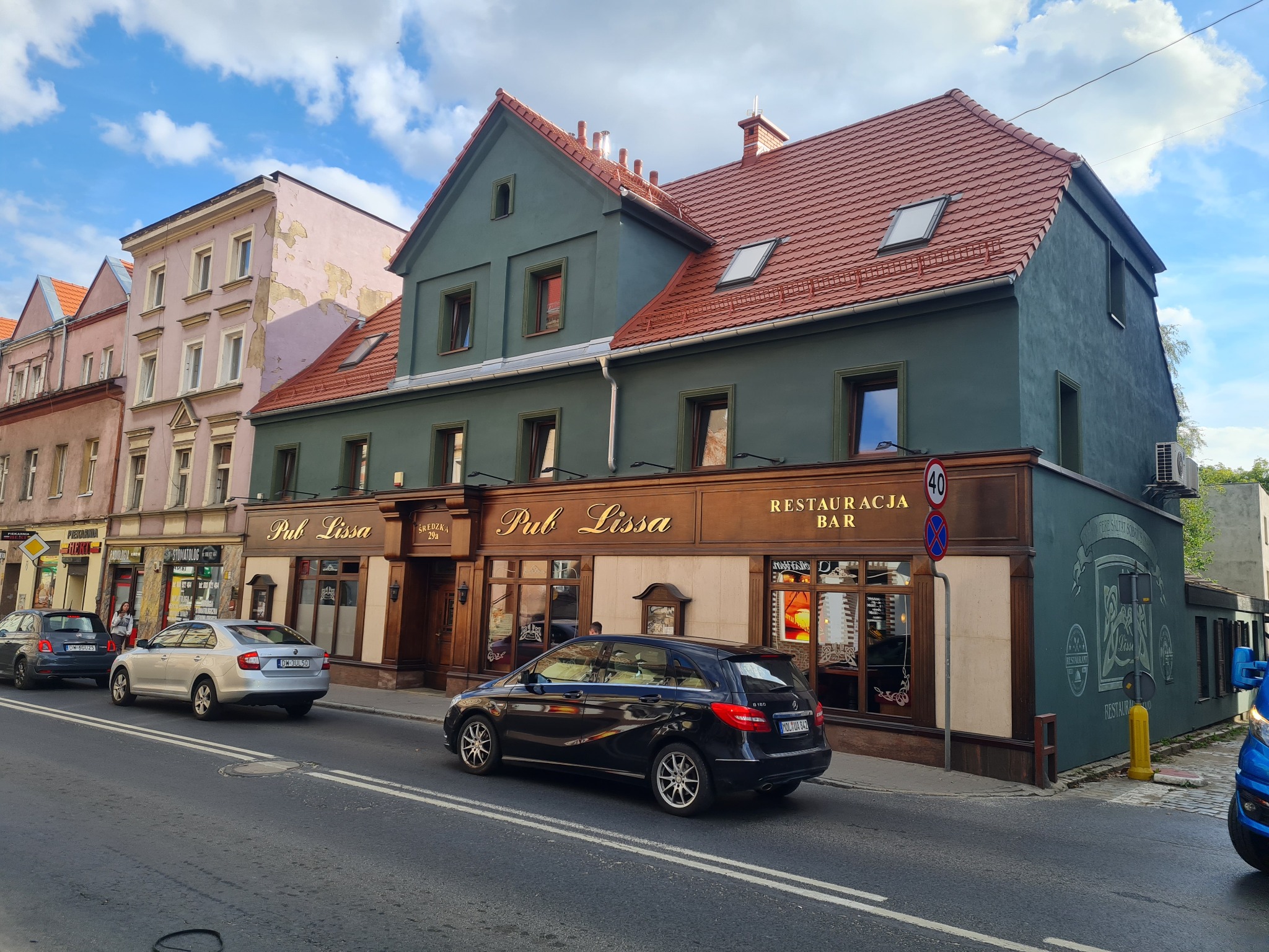 Odnowiona fasada kamienicy z szyldem 'Pub Lissa' i 'Restauracja Bar', z nową elewacją w kolorze zieleni i brązu, widok z ulicy, samochody na pierwszym planie.