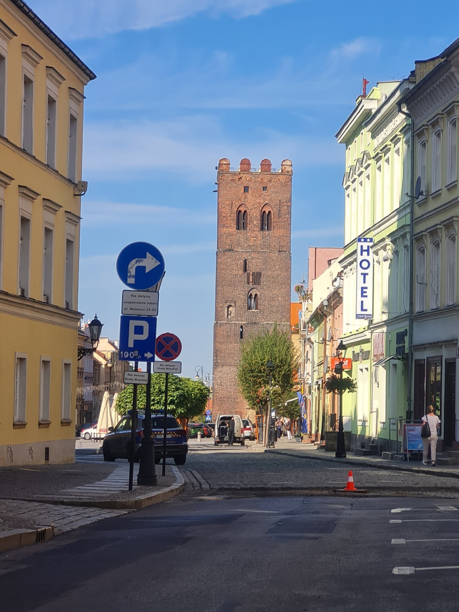 Ulica w mieście z wysoką, ceglaną wieżą w tle. Budynki po bokach ulicy w pastelowych kolorach, znaki drogowe i pojedyncze osoby na chodniku.