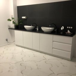 Piotr Ciołek - Nowoczesna łazienka z dwoma owalnymi umywalkami nablatowymi, czarną armaturą, białymi szafkami pod umywalkami i czarnymi płytkami na ścianie, uzupełniona rośliną w doniczce i podświetlanym...