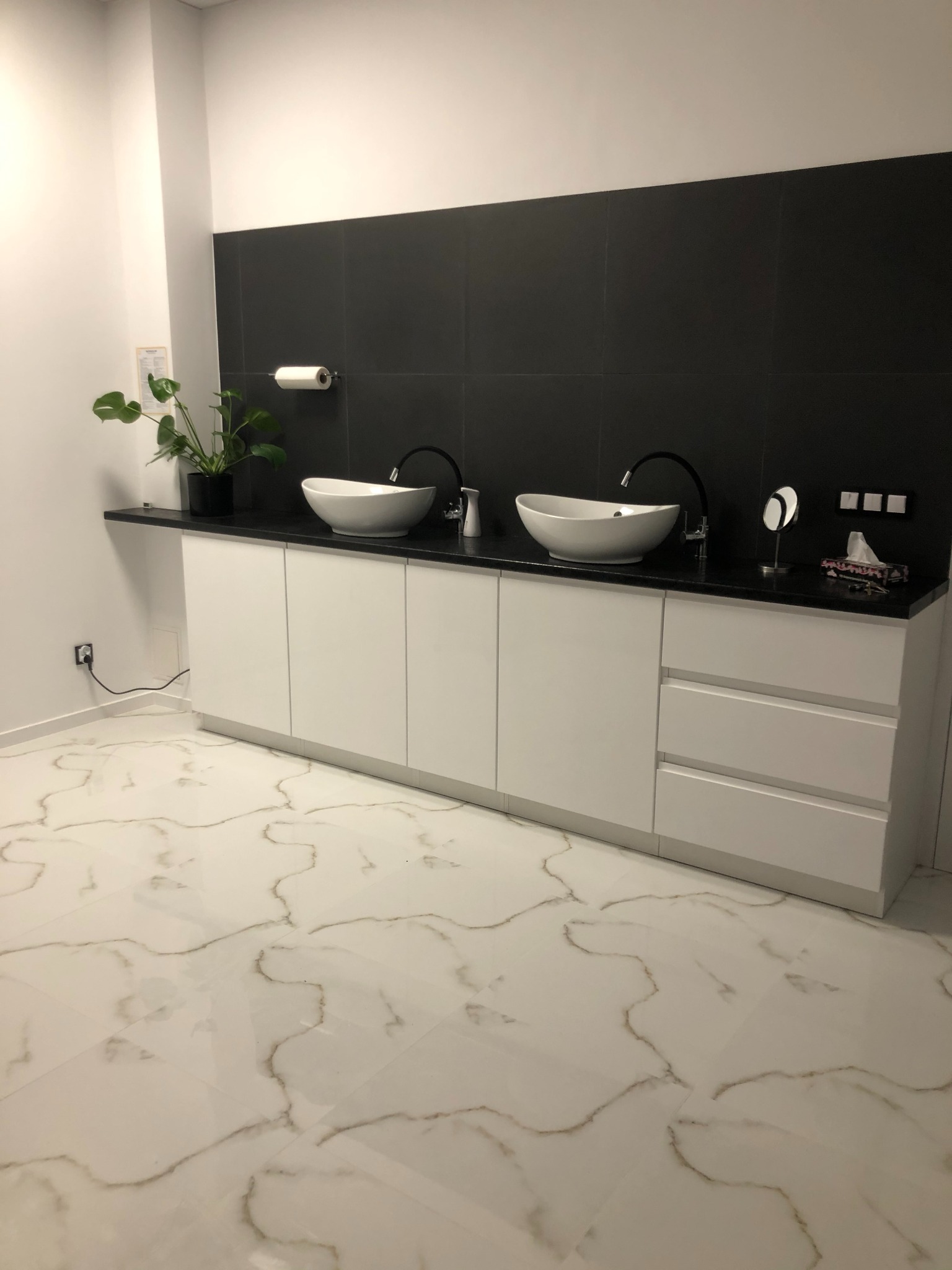 Nowoczesna łazienka z dwoma owalnymi umywalkami nablatowymi, czarną armaturą, białymi szafkami pod umywalkami i czarnymi płytkami na ścianie, uzupełniona rośliną w doniczce i podświetlanym...