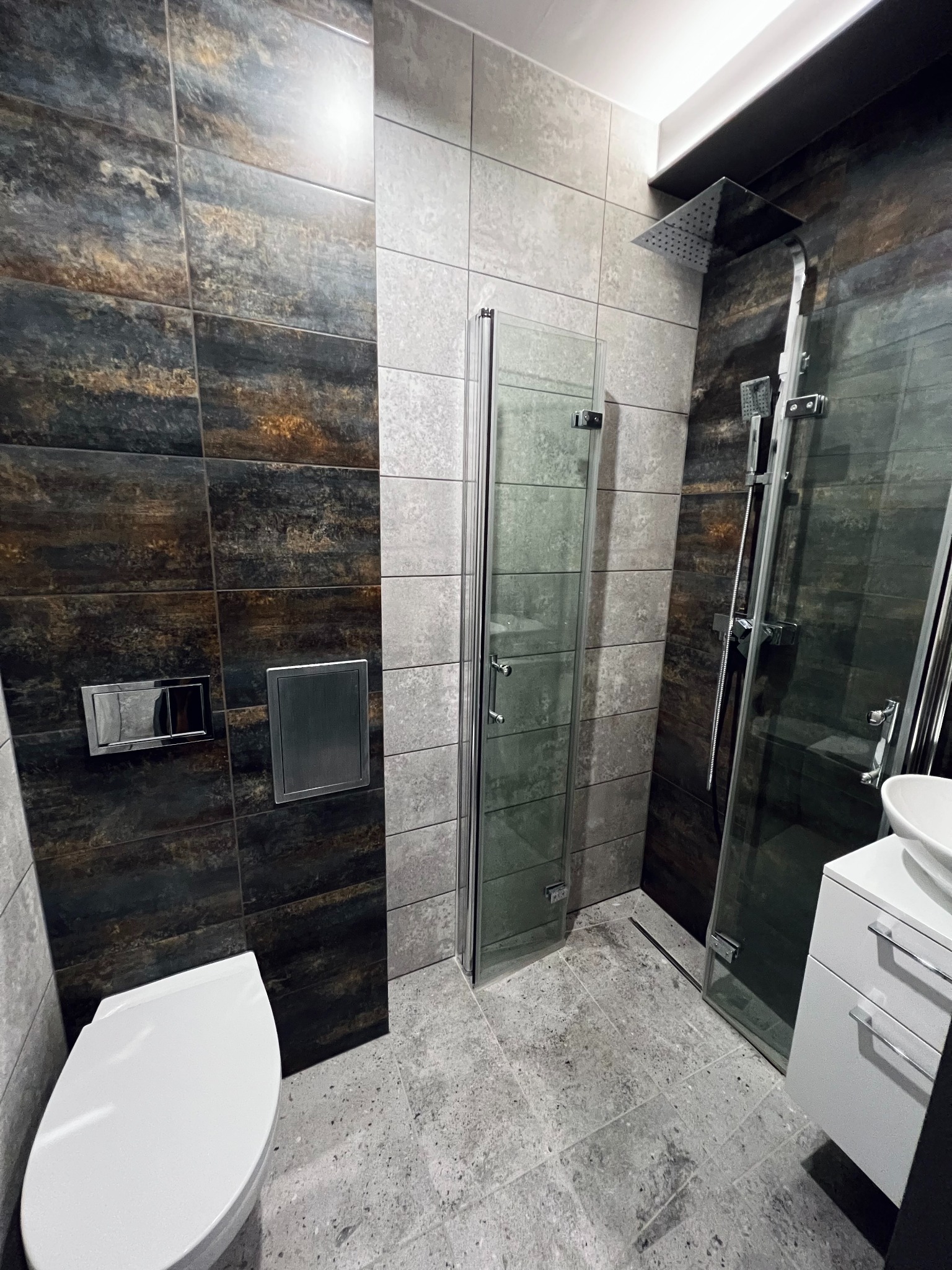 Nowoczesna łazienka z kabiną prysznicową typu walk-in, toaletą podwieszaną z chromowanym przyciskiem spłukiwania, umywalką nablatową i płytkami imitującymi metal na ścianach.