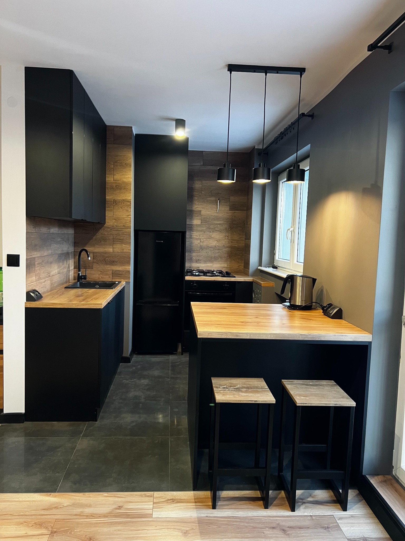 Nowoczesna kuchnia w ciemnych barwach z drewnianymi akcentami: czarne szafki, drewnopodobna okładzina ściany, drewniany blat, czarne hokery z drewnianymi siedziskami, wiszące lampy nad wyspą.