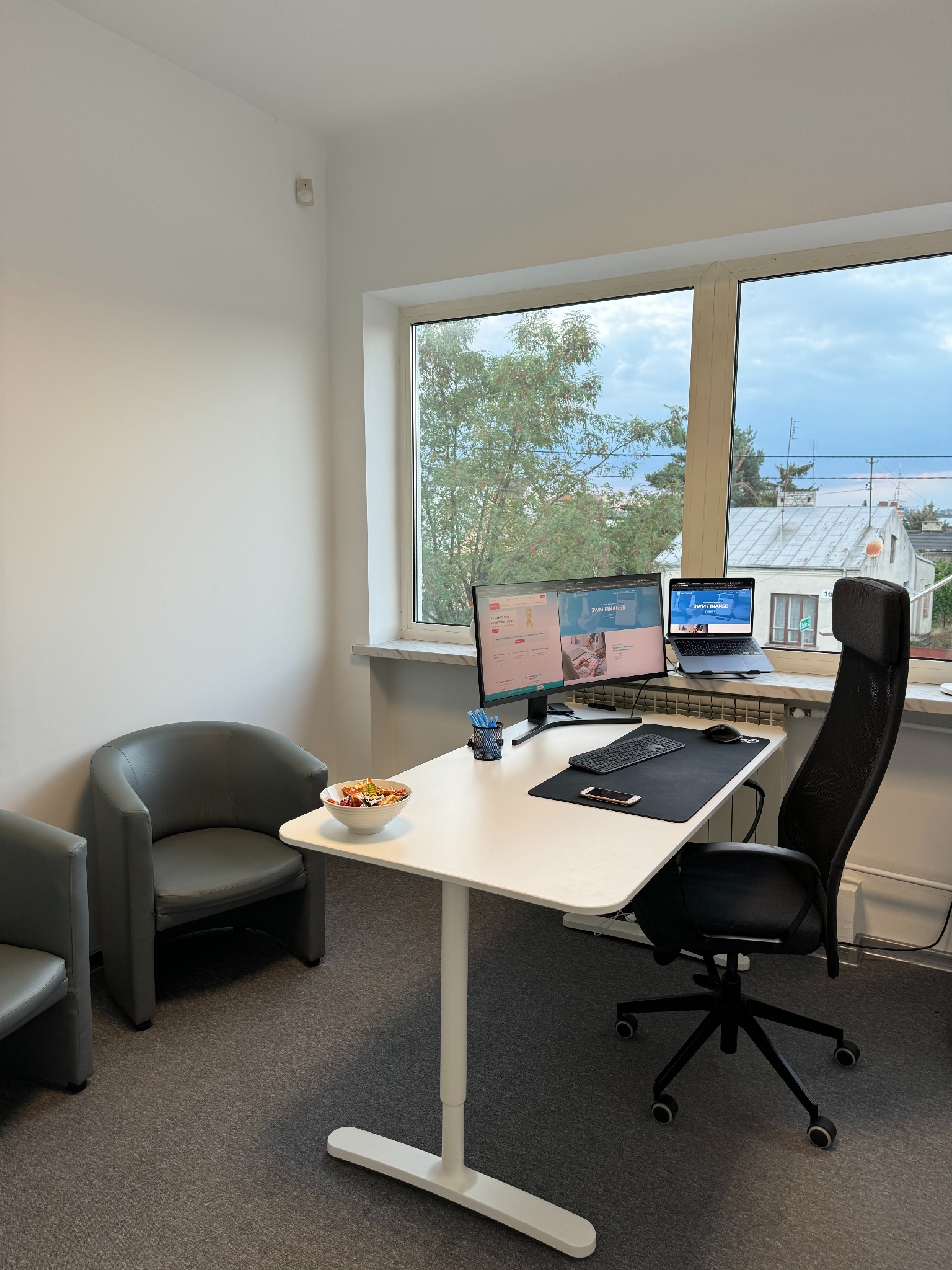 Jasne biuro z białym biurkiem, dwoma monitorami, laptopem, miską owoców i widokiem na zewnątrz przez okno, sugerujące nowoczesne środowisko pracy.