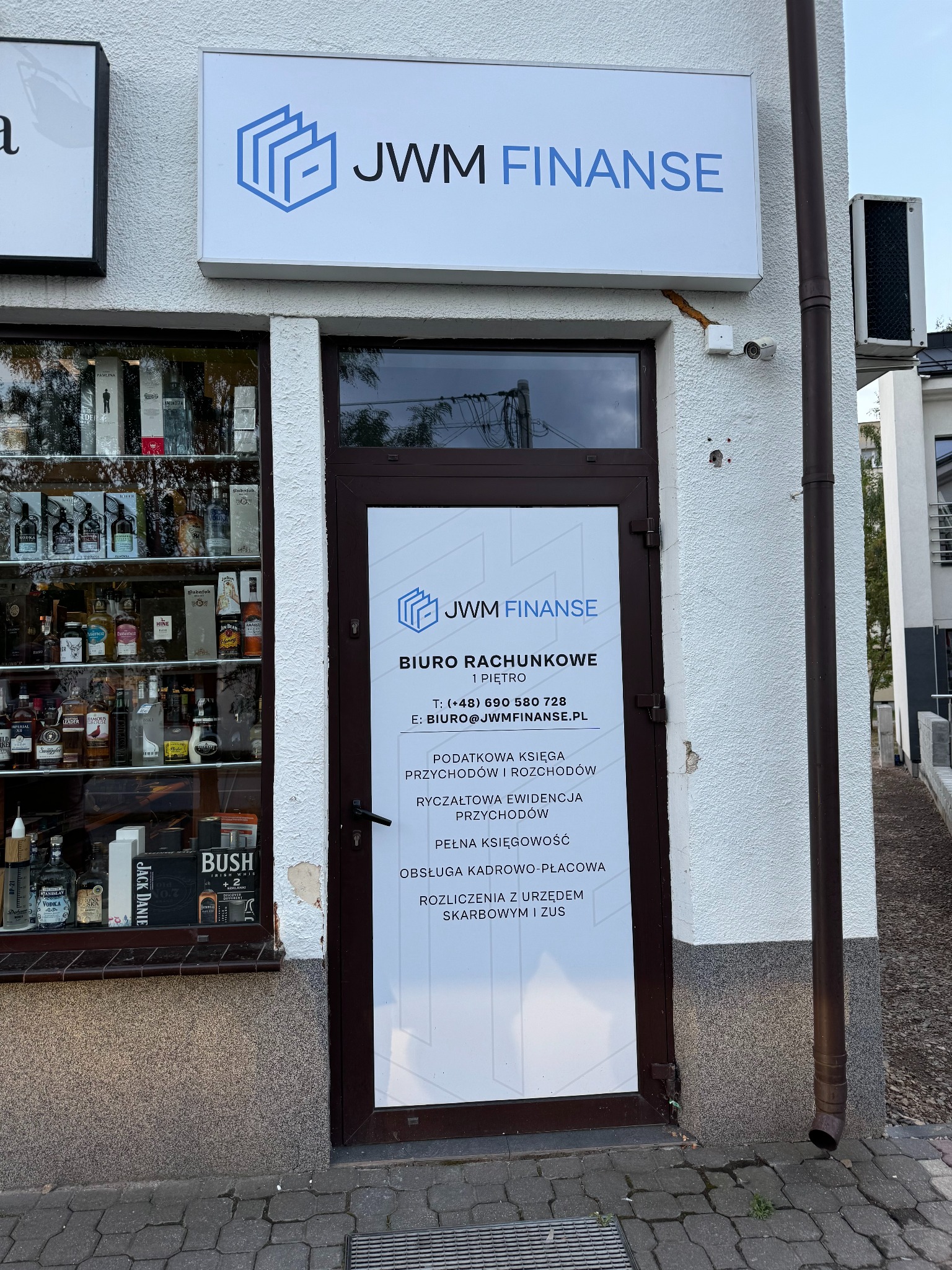 Wejście do biura rachunkowego JWM Finanse z widocznym logo i informacjami kontaktowymi na drzwiach, obok witryna z ekspozycją alkoholi.