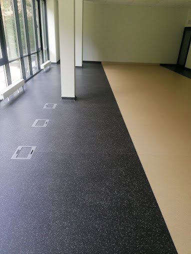 Częściowo wykończone wnętrze z podłogą podzieloną na dwie sekcje: ciemnoszarą, prawdopodobnie gumową wykładzinę z wbudowanymi gniazdami podłogowymi i jasnobeżową, przygotowaną pod dalsze prace...