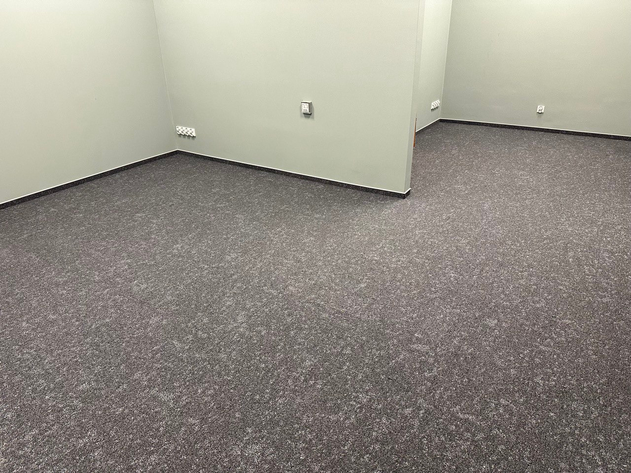 Szare płytki dywanowe ułożone w dwóch pustych pokojach biurowych z jasnymi ścianami, widoczne gniazdka elektryczne.