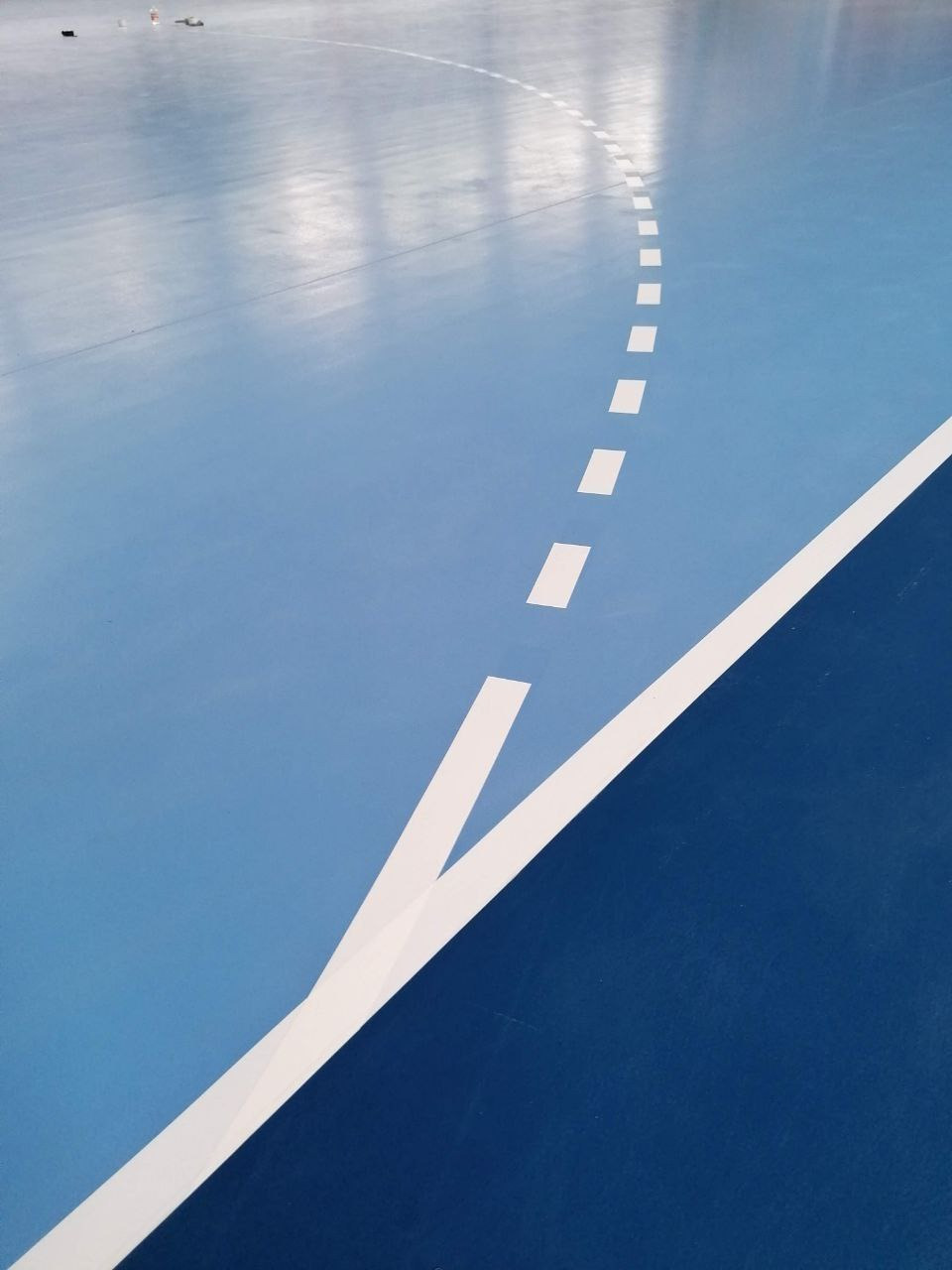 Niebieska podłoga hali sportowej z białymi liniami wyznaczającymi pole gry, widoczny fragment linii rzutów karnych i linii bocznej.