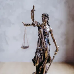 Brązowa statuetka Temidy, bogini sprawiedliwości, z wagą i mieczem, stojąca na drewnianym blacie na tle szarej ściany.