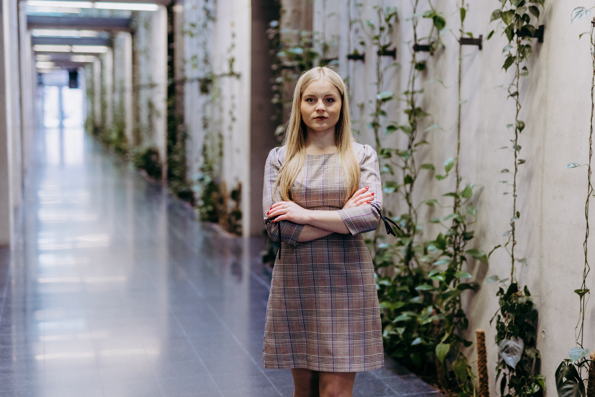 Portret kobiety z blond włosami i w kraciastej sukience stojącej na tle korytarza z roślinami pnącymi się po ścianach.