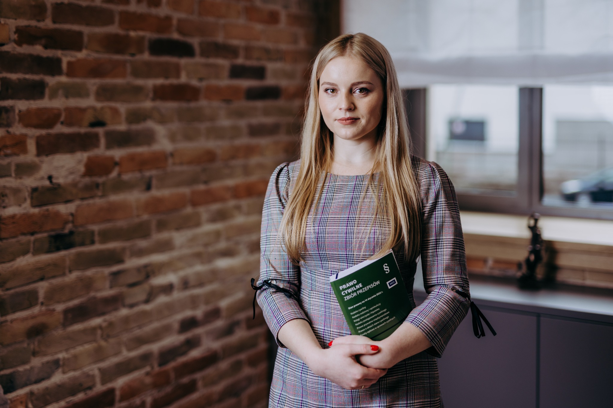 Portret kobiety trzymającej zielony kodeks 'Prawo Cywilne' na tle ceglanej ściany i okna.