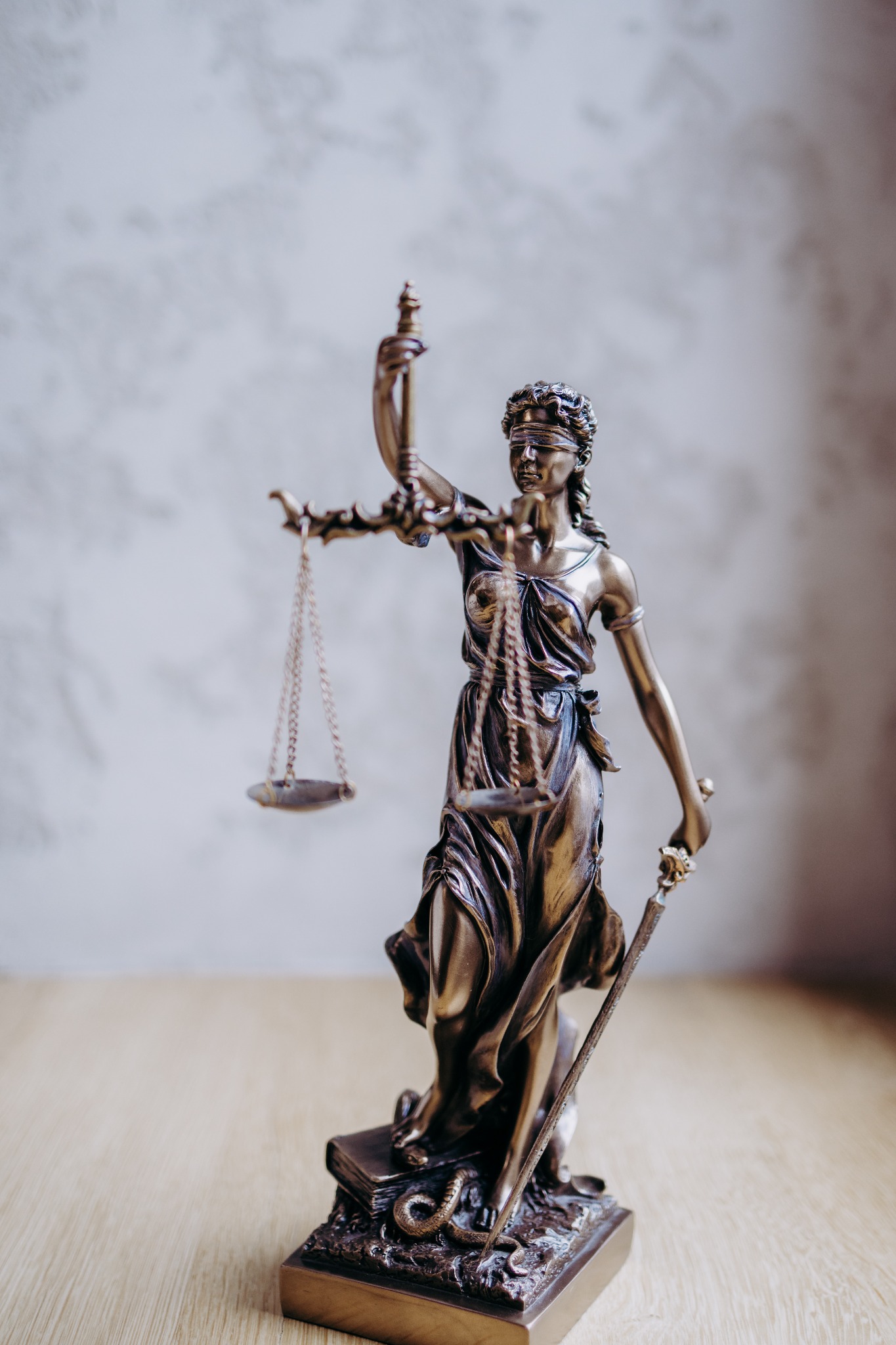 Brązowa statuetka Temidy, bogini sprawiedliwości, z wagą i mieczem, stojąca na drewnianym blacie na tle szarej ściany.