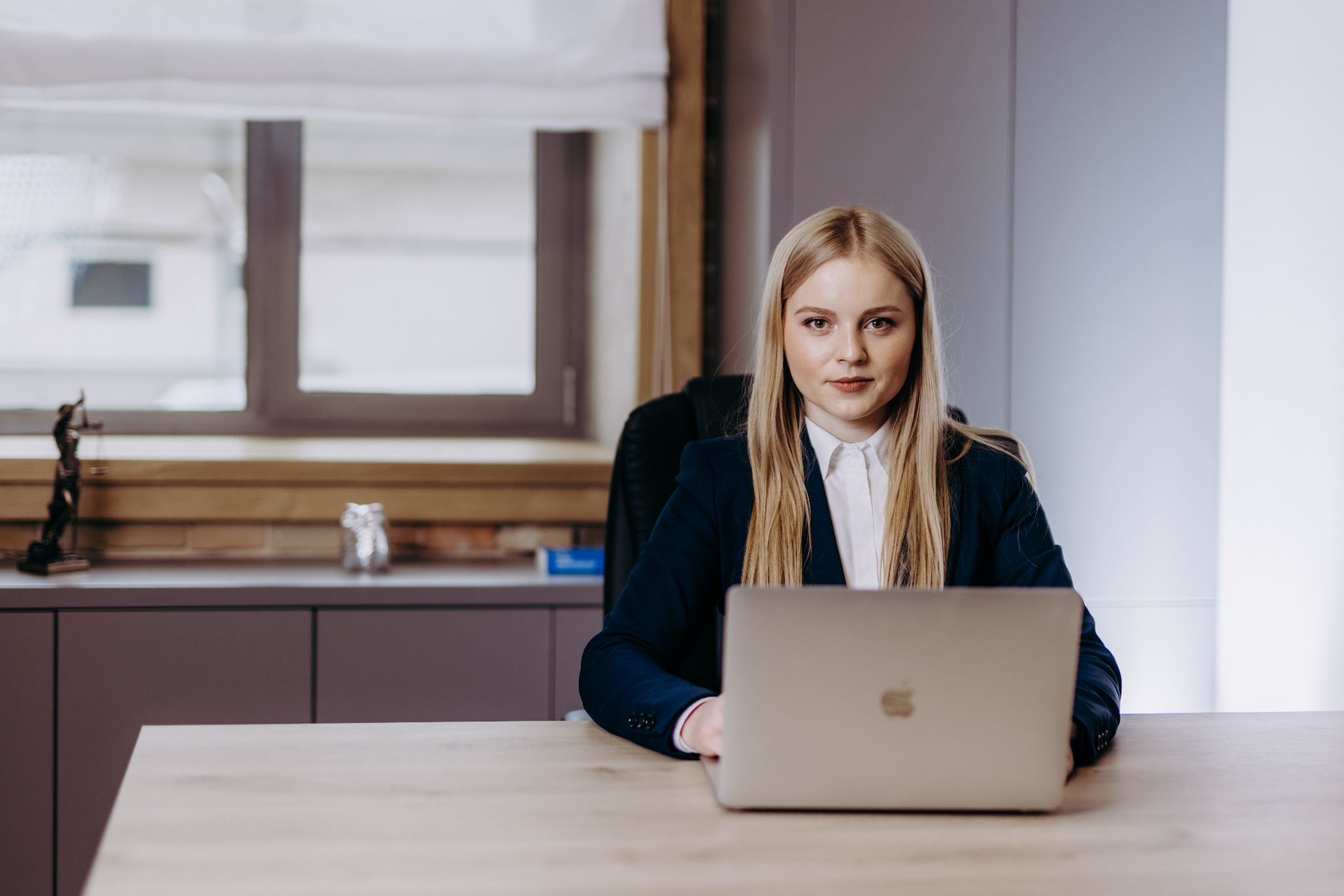 Portret kobiety w biurze, w tle statuetka Temidy i okno, kobieta w marynarce pracuje na laptopie.