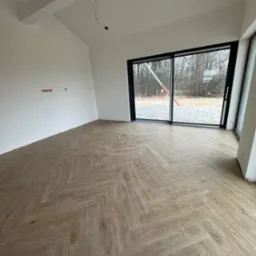 Jasne panele ułożone w jodełkę w przestronnym, surowym wnętrzu z dużymi przesuwnymi oknami i widocznymi instalacjami elektrycznymi.