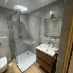 Nowoczesna łazienka w szarości z drewnianymi akcentami: kabina prysznicowa ze szklanymi ścianami, umywalka z szafką i lustro z oświetleniem LED.