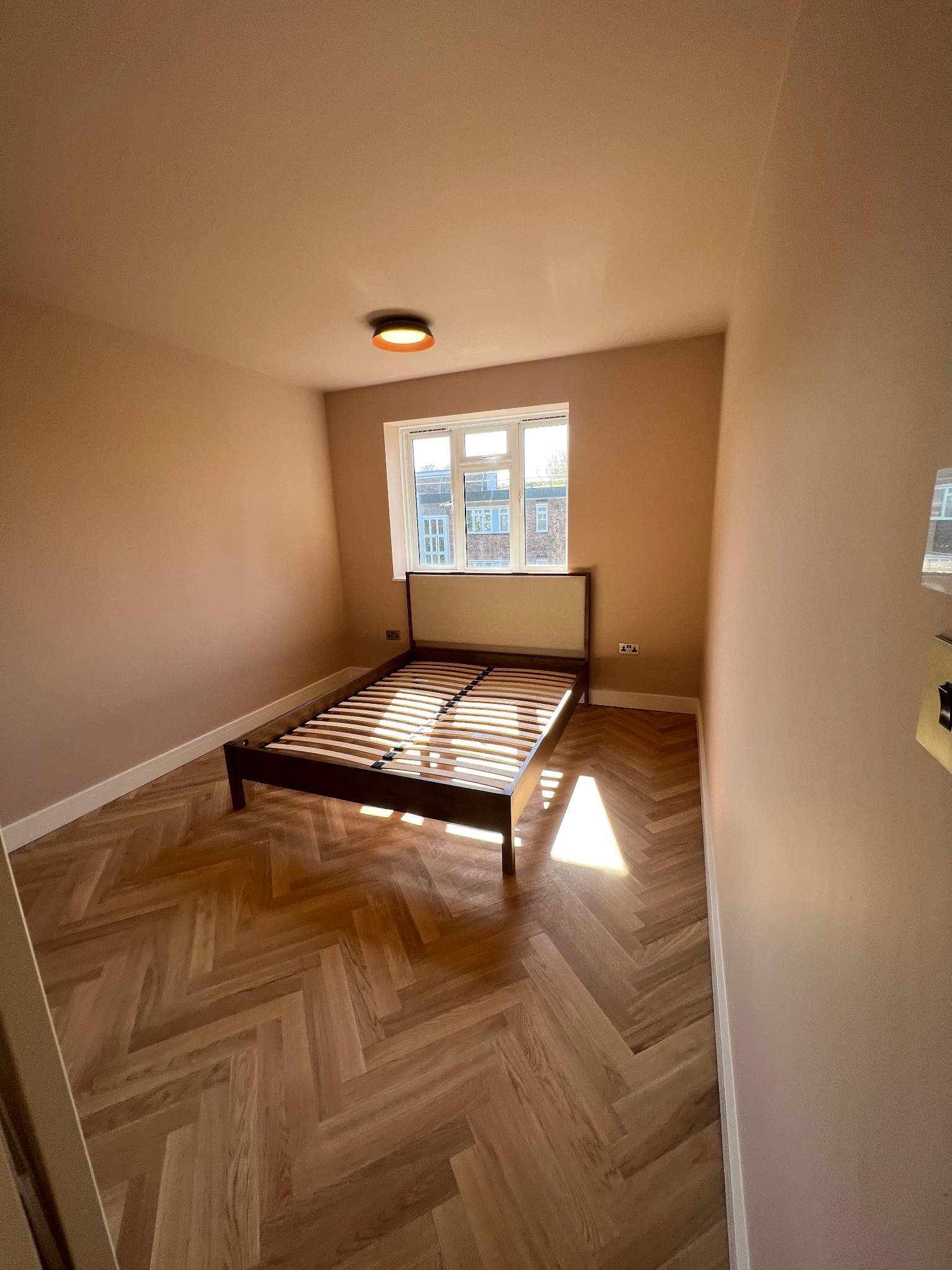 Pokój z parkietem ułożonym we wzór jodełki, świeżo pomalowanymi ścianami w odcieniu pudrowego różu i minimalistycznym łóżkiem bez materaca, oświetlony naturalnym światłem wpadającym przez okno.