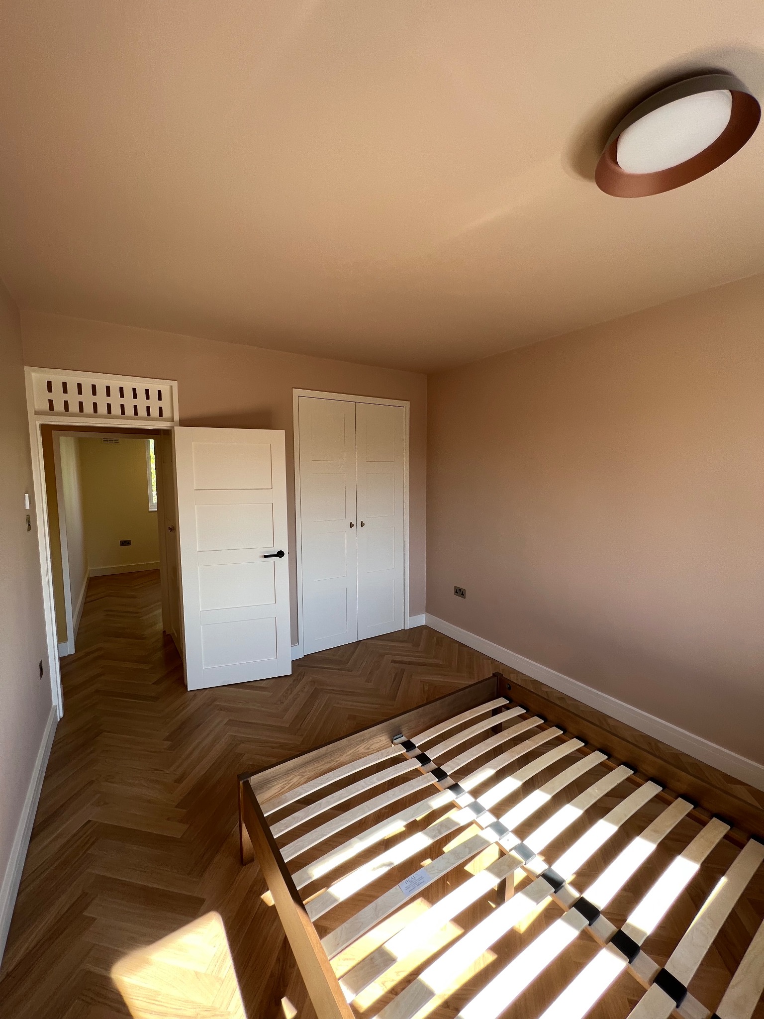 Pokój z parkietem ułożonym w jodełkę, pomalowanymi na beżowo ścianami, białymi drzwiami z czarną klamką i szafą wnękową, oraz drewnianym stelażem łóżka bez materaca wpadającym w promienie słońca.