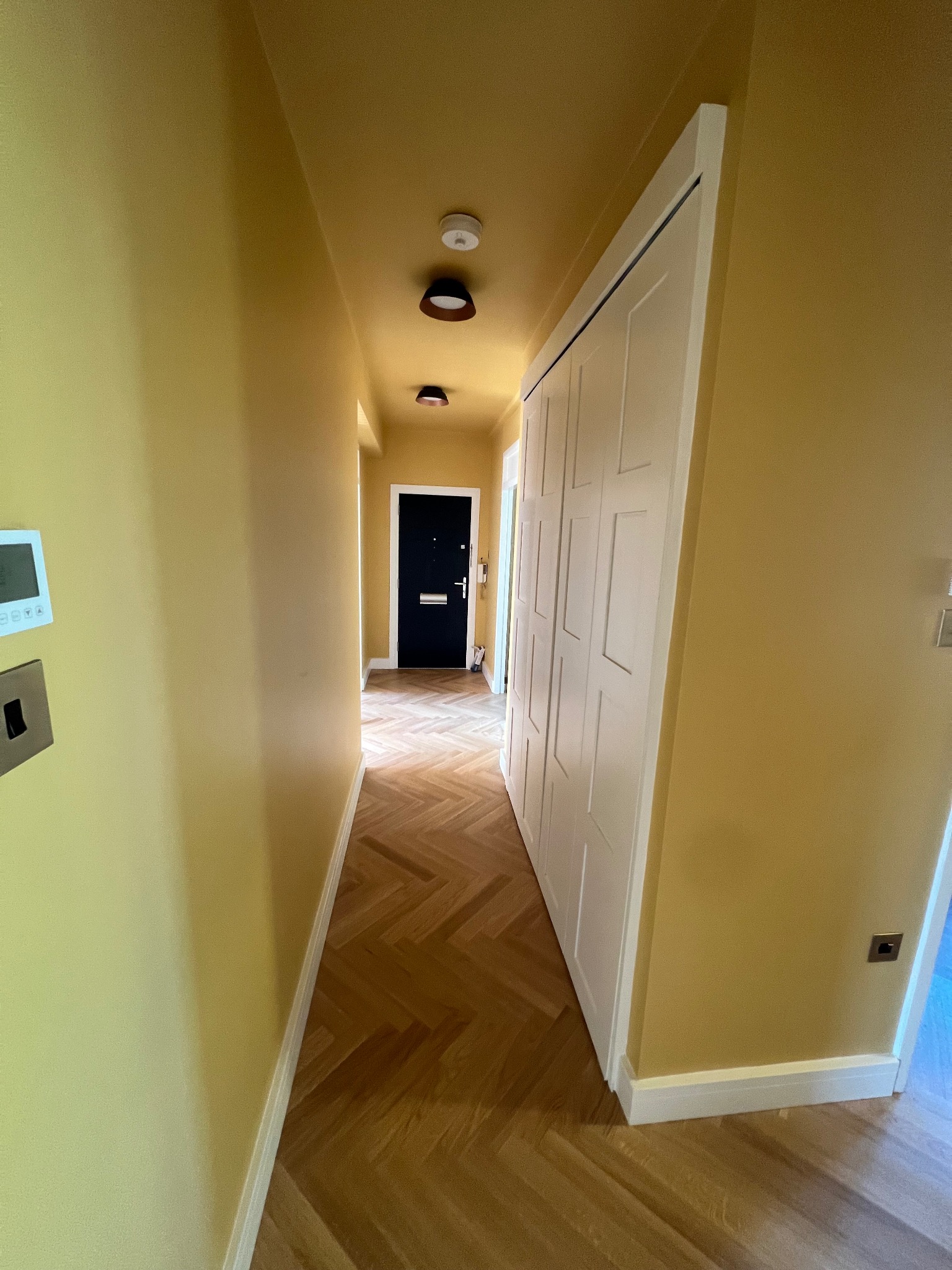 Korytarz po remoncie z parkietem ułożonym w jodełkę, pomalowanymi na żółto ścianami i sufitem, oraz białą zabudową szafy wnękowej z przesuwnymi drzwiami.