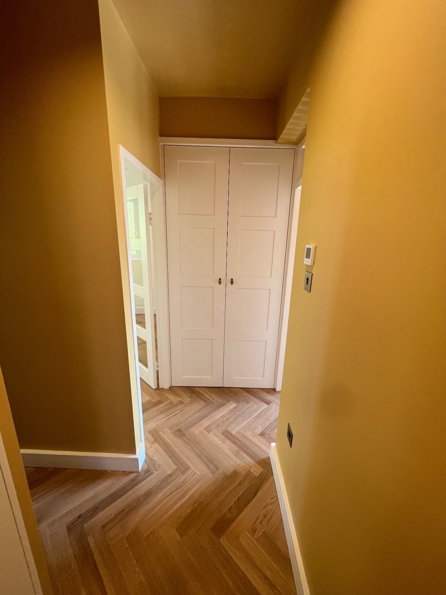 Korytarz z parkietem ułożonym w jodełkę, pomalowany na odcień musztardowy, z białymi drzwiami i listwami przypodłogowymi.