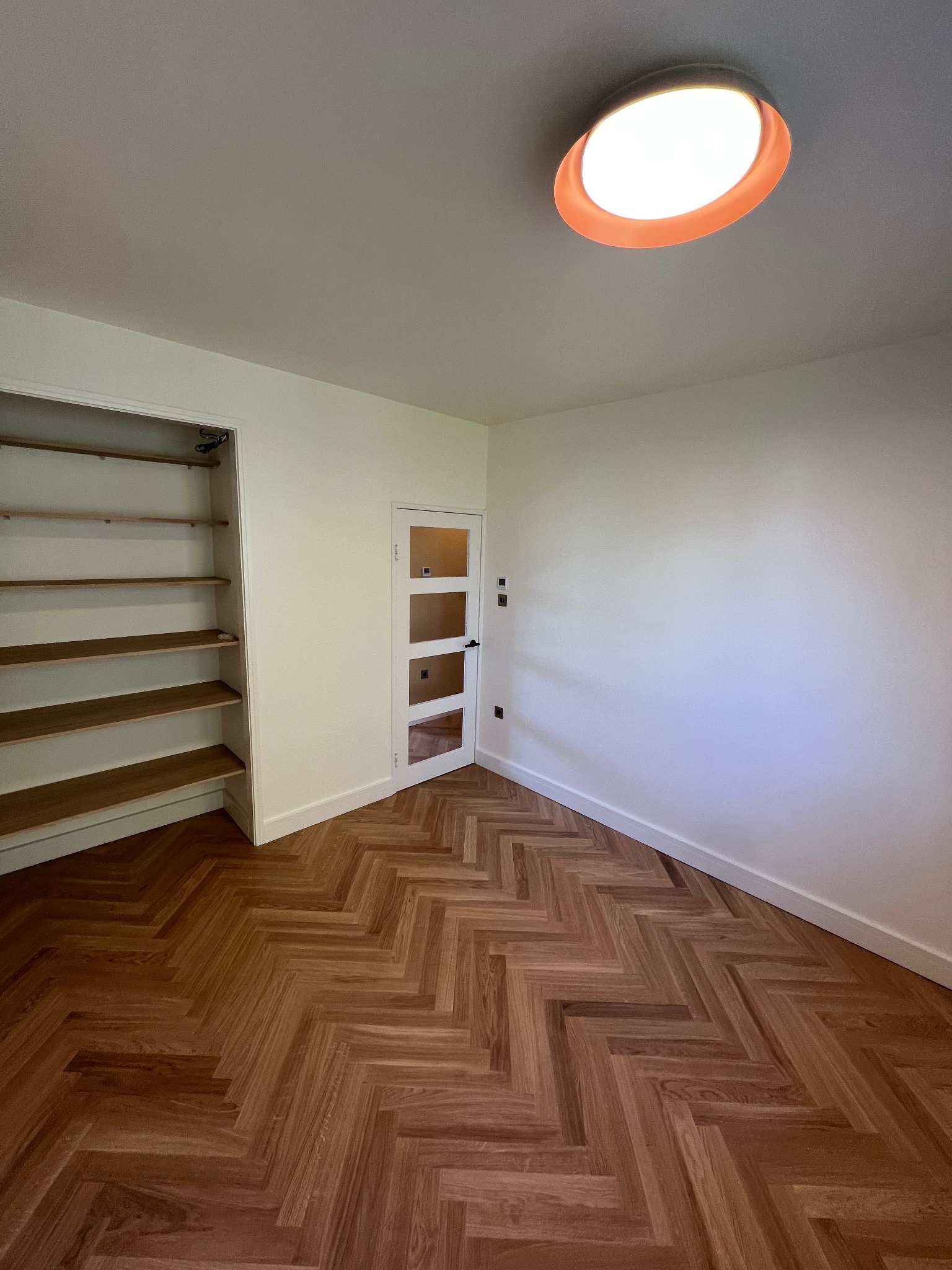 Wnętrze pokoju z parkietem ułożonym w jodełkę, białe ściany, wbudowana szafa z drewnianymi półkami i przeszklone drzwi z czarną klamką, okrągła lampa sufitowa z pomarańczową obwódką.