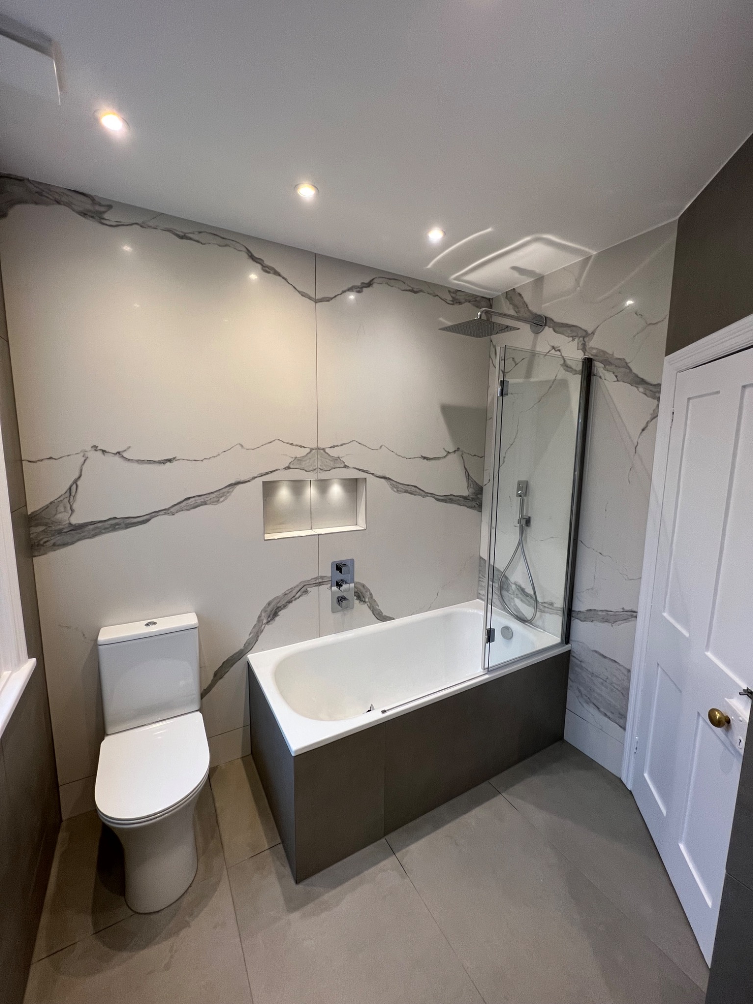 Nowoczesna łazienka z toaletą, wanną z parawanem, płytkami imitującymi marmur i wnęką z oświetleniem punktowym.