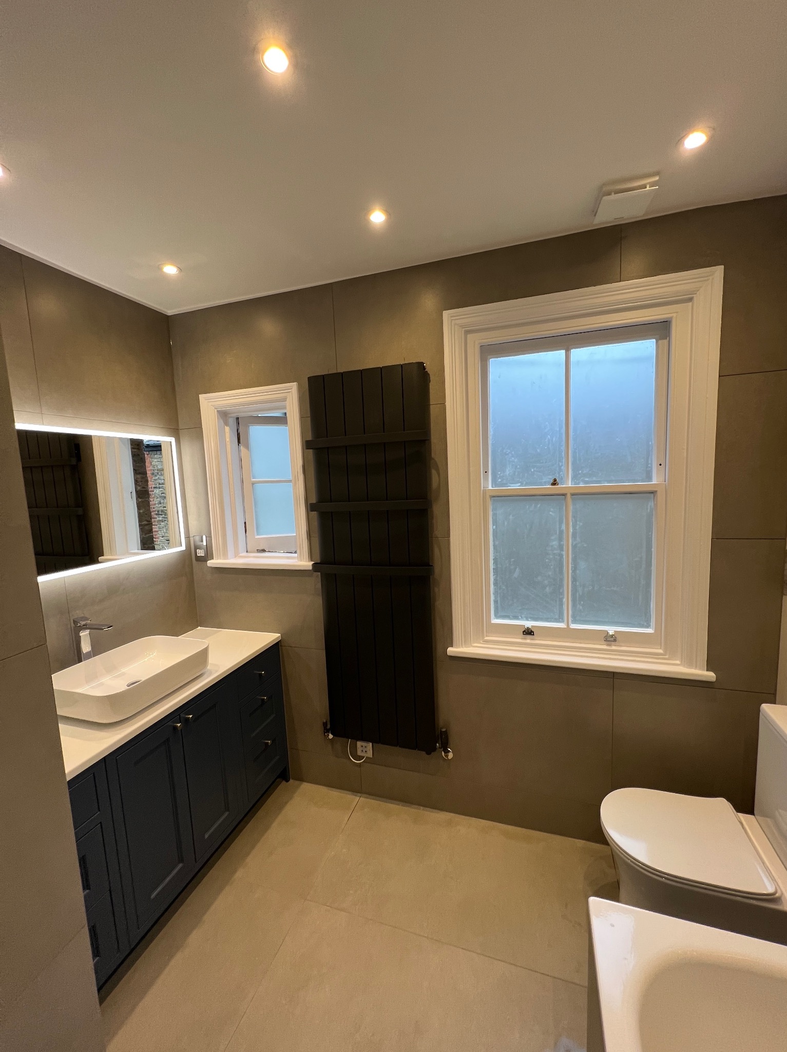 Nowoczesna łazienka z szarymi płytkami, podświetlanym lustrem nad białą umywalką nablatową osadzoną na ciemnogranatowej szafce, czarnym grzejnikiem drabinkowym i białymi oknami skrzynkowymi.