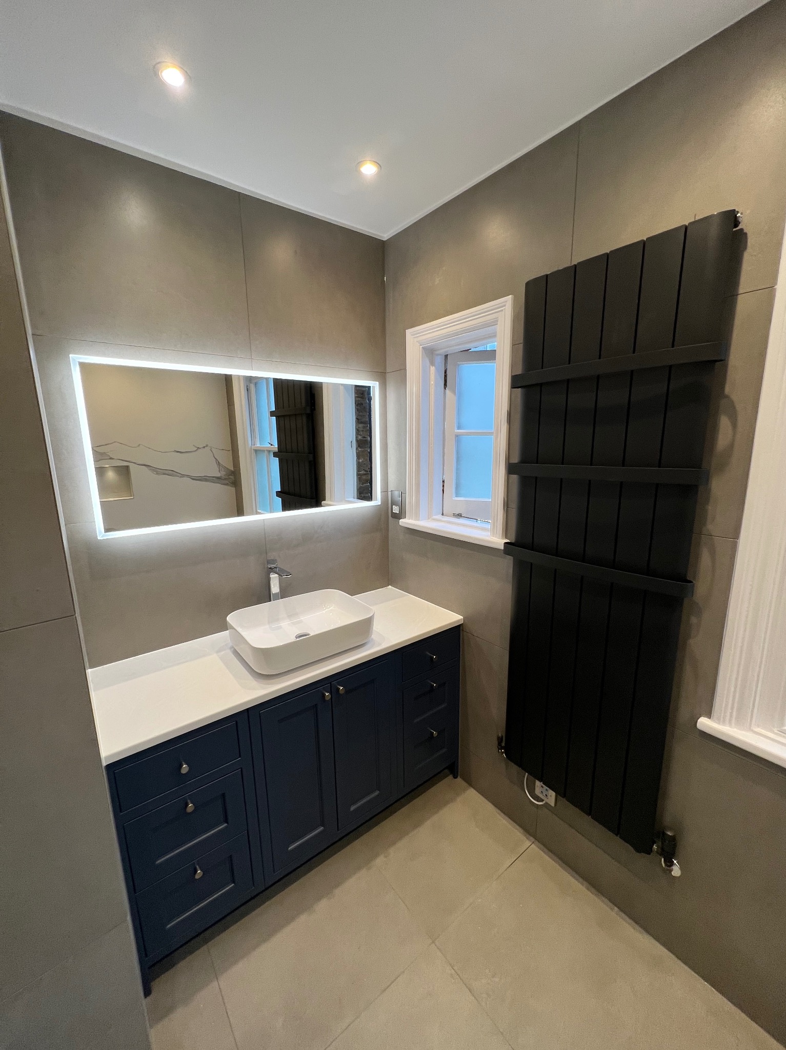 Wykończona łazienka z szarymi płytkami, zabudową umywalkową w kolorze granatowym z białym blatem, prostokątną umywalką nablatową, podświetlanym lustrem, czarnym grzejnikiem drabinkowym i oknem...