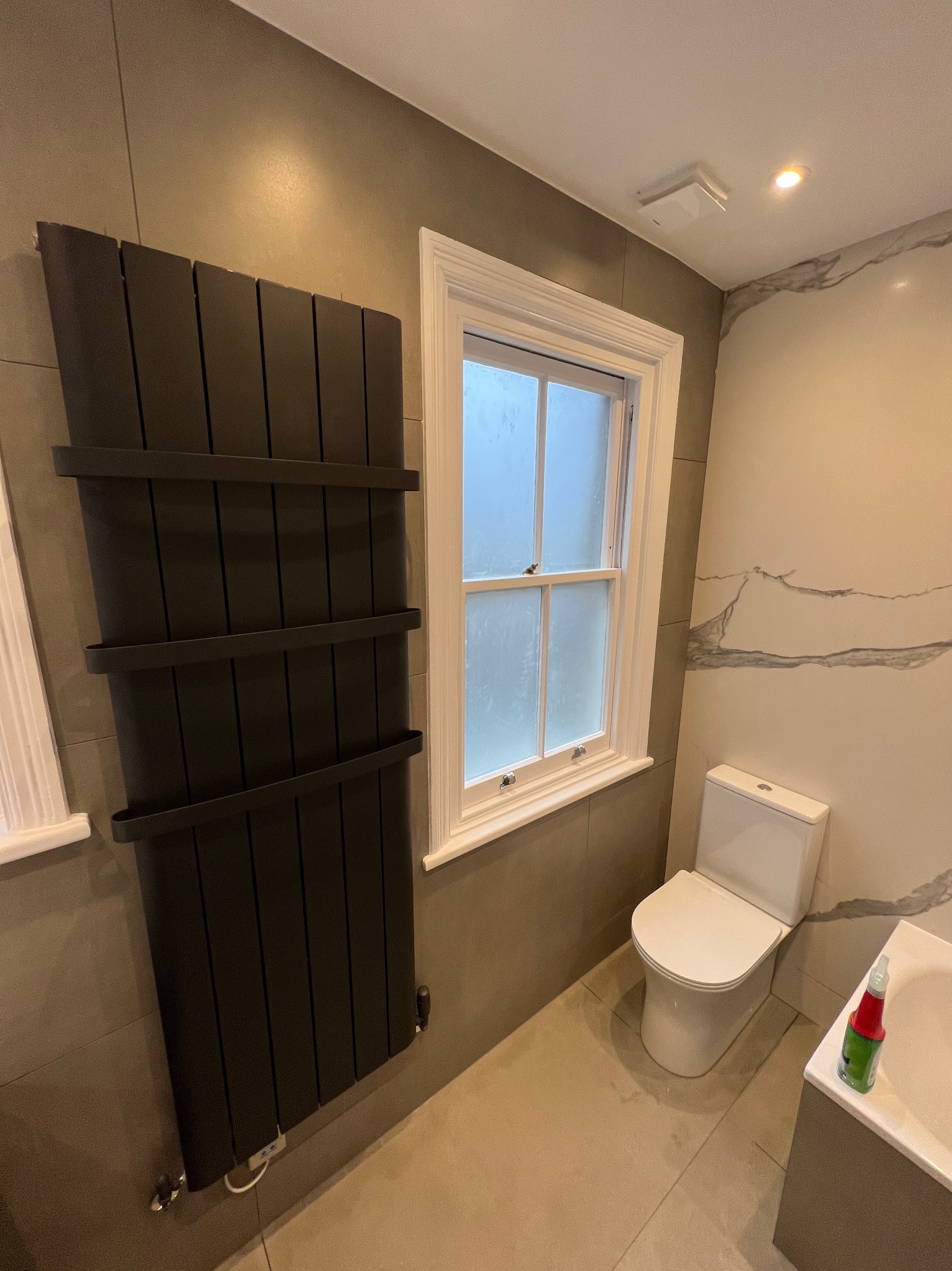 Wykończona łazienka w szarościach z grzejnikiem drabinkowym, toaletą i oknem z białymi ramami, marmurowe płytki na ścianie.