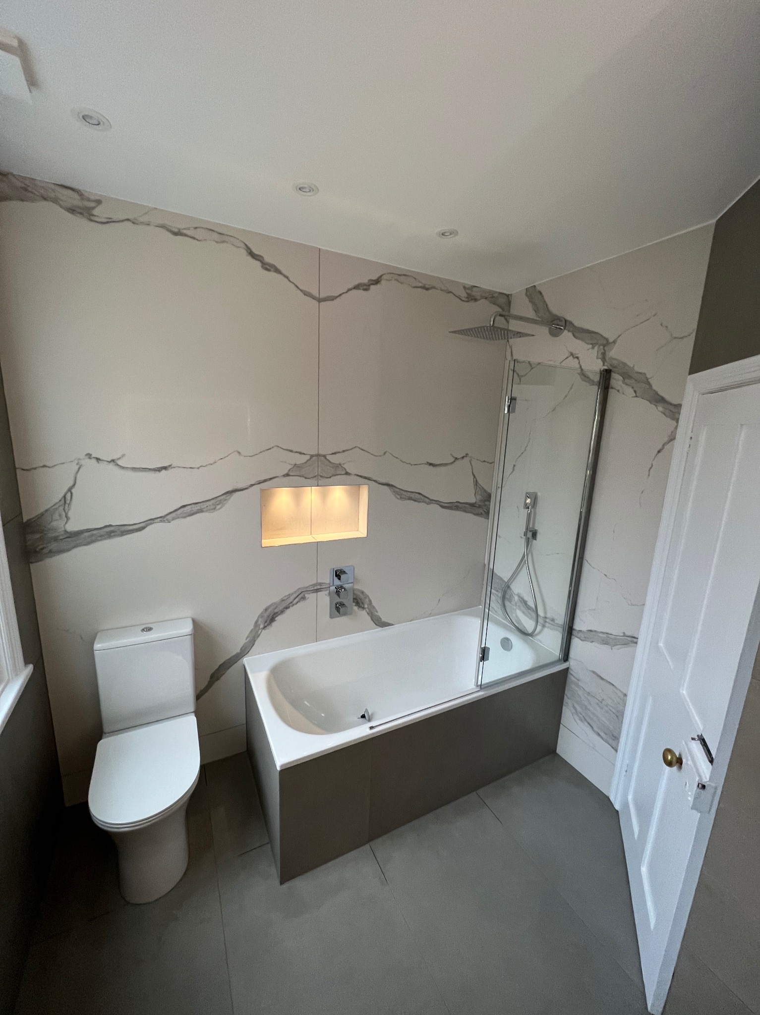 Wykończona łazienka z wanną z szarym obramowaniem, toaletą i kabiną prysznicową, ściany wyłożone płytkami imitującymi marmur z podświetlaną wnęką i szarą podłogą.