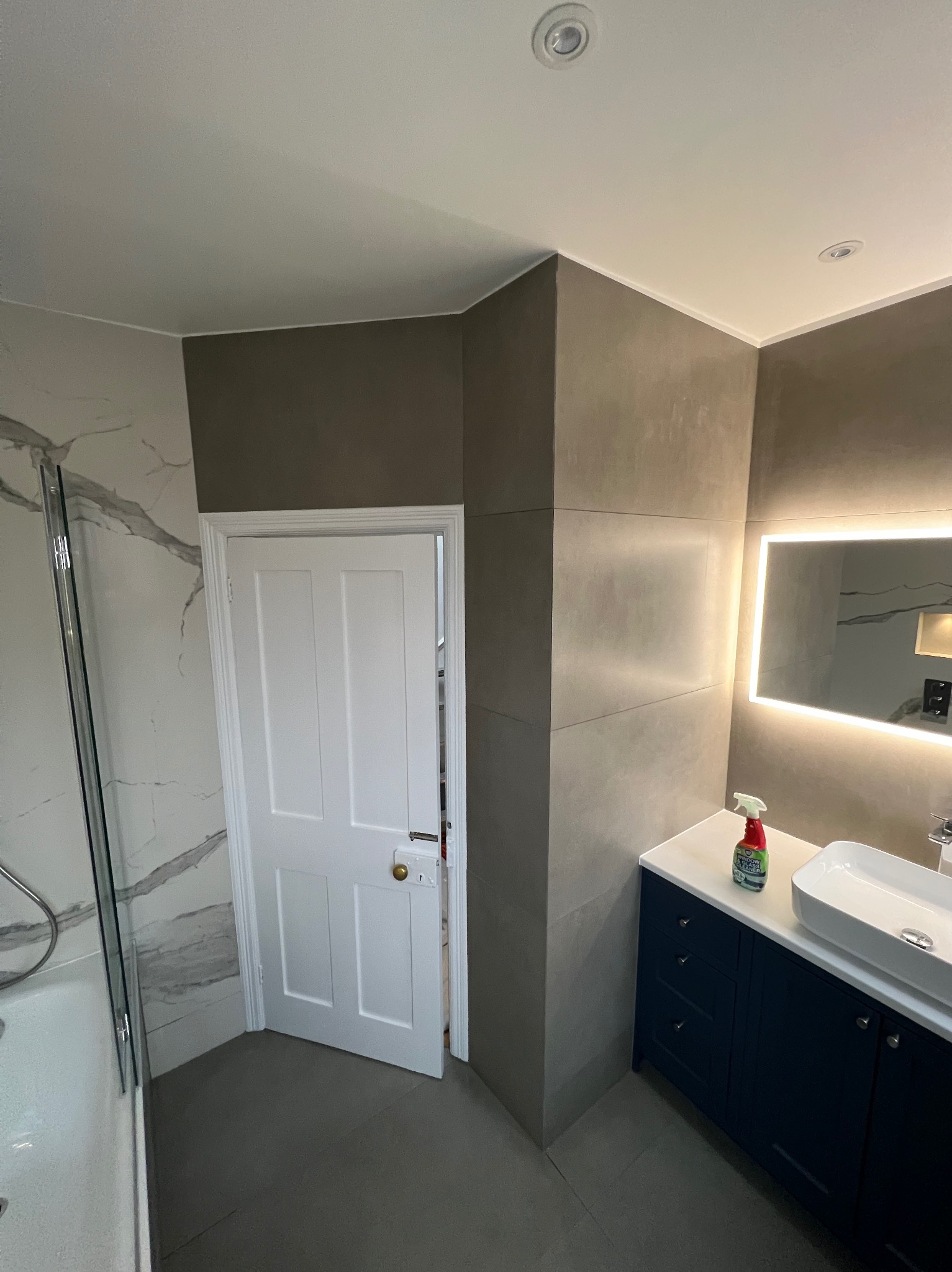 Nowoczesna łazienka z marmurowymi płytkami pod prysznicem, szarobrązowymi panelami ściennymi, białą armaturą i granatową szafką pod umywalką, oświetlonym lustrem oraz otwartymi białymi drzwiami.